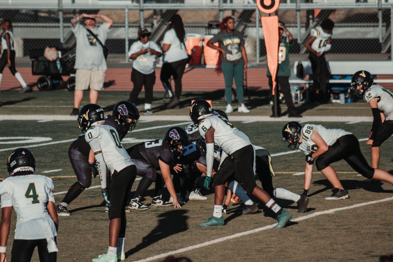 High school football teams line up at scrimmage during an evening game with spectators watching from sidelines.