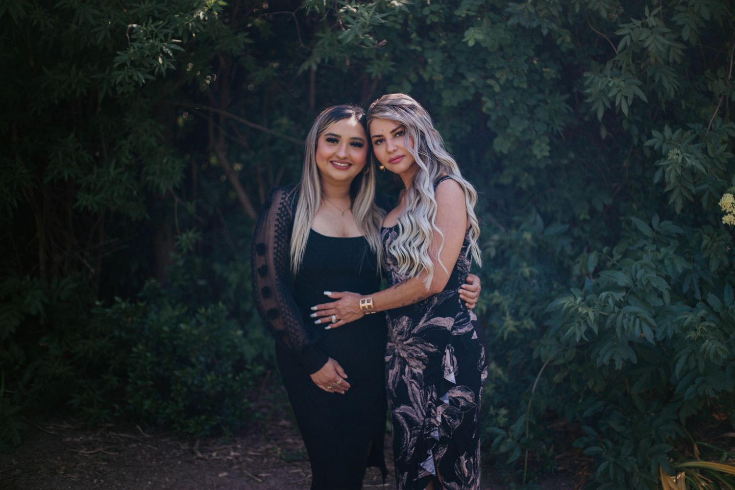 Two people in dark clothing pose together outdoors against a leafy green background at dusk.