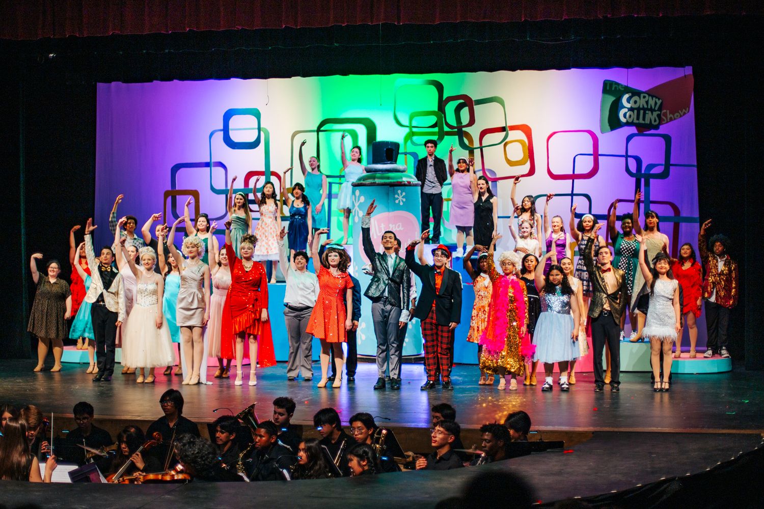 Performers on stage in colorful costumes stand together for finale with vibrant geometric backdrop and orchestra pit below.