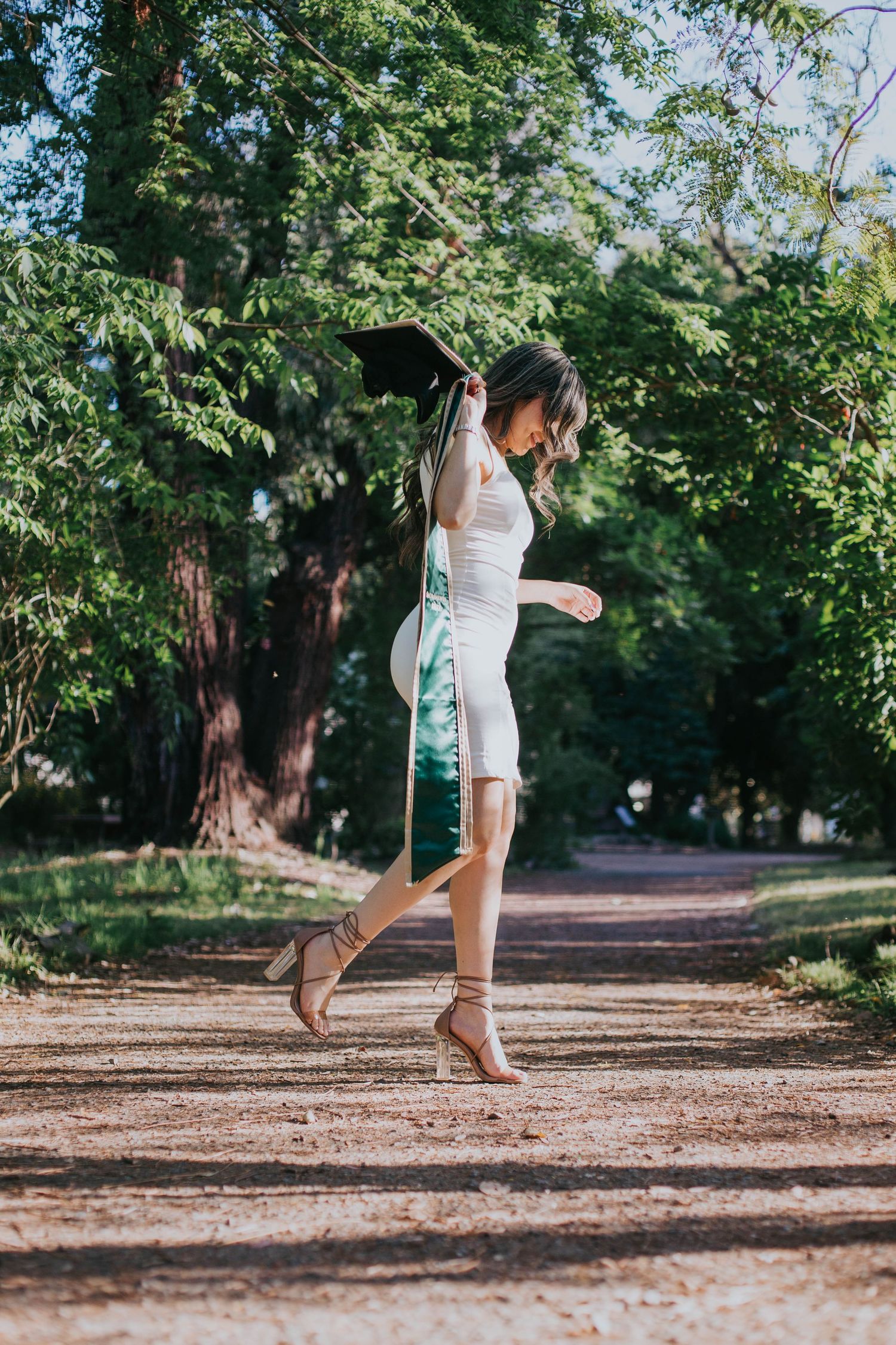 A person in a white dress and brown boots jumps joyfully on a tree-lined dirt path on a sunny summer day.