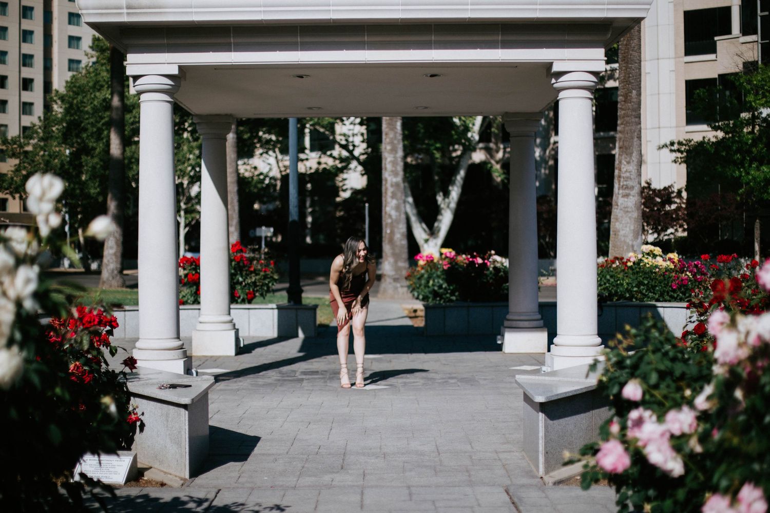 A person stands beneath a white columned gazebo with blooming flowers in the foreground and architecture in the background.