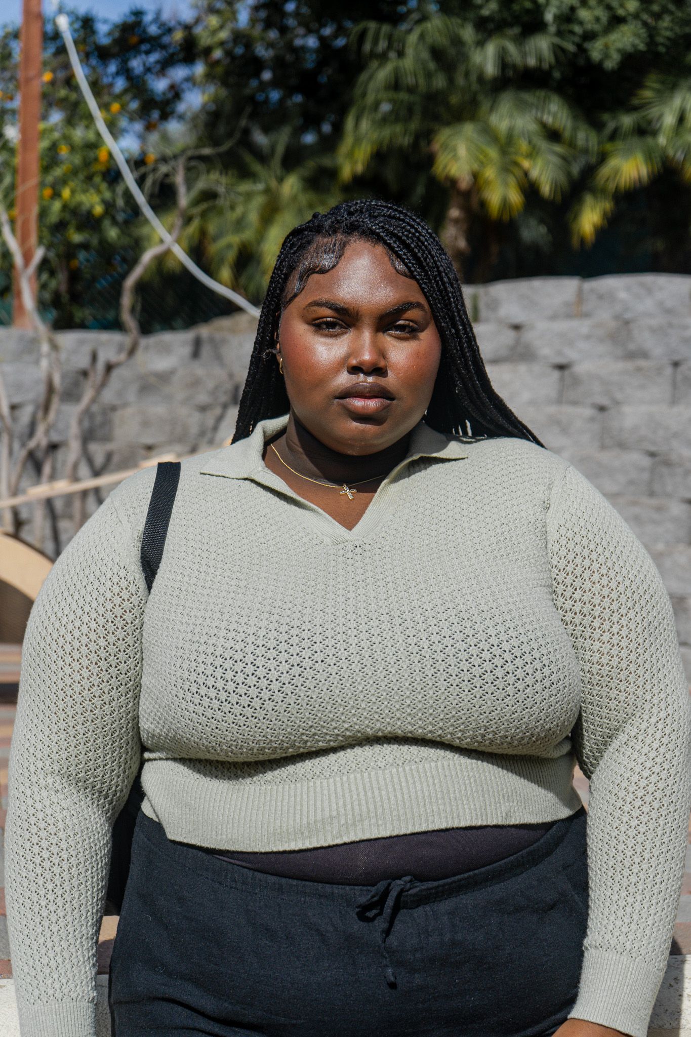 A person wearing a cream knit sweater and dark pants stands outdoors against a stone wall with palm trees in background.