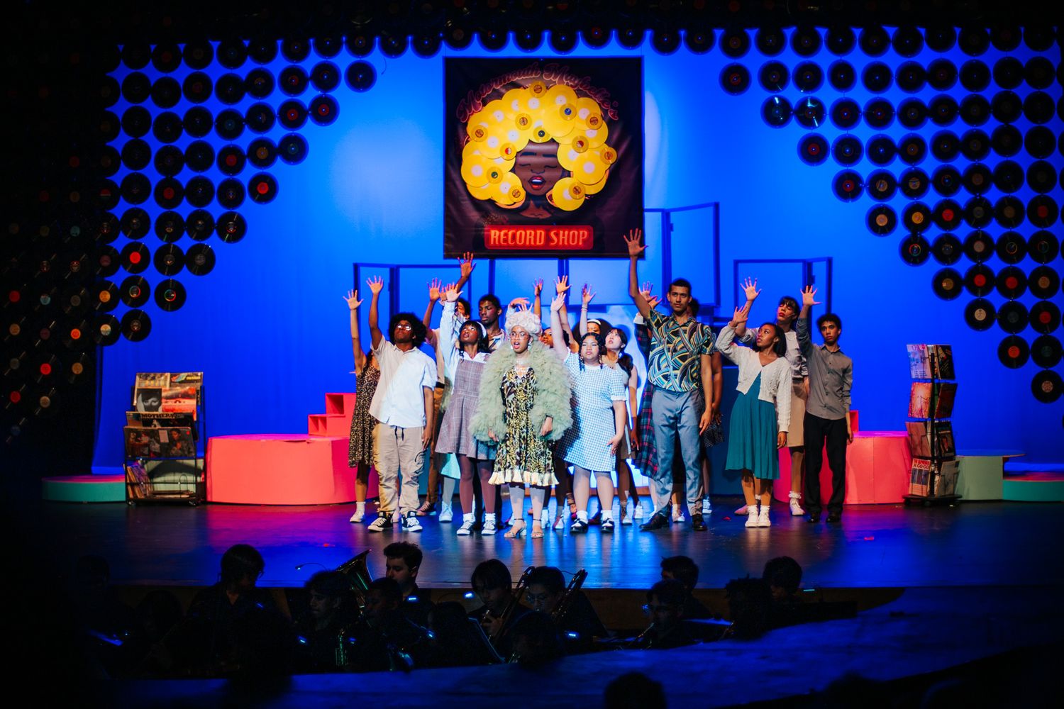 Performers stand on a colorful stage with blue lighting and a large circular logo display during a theatrical production.