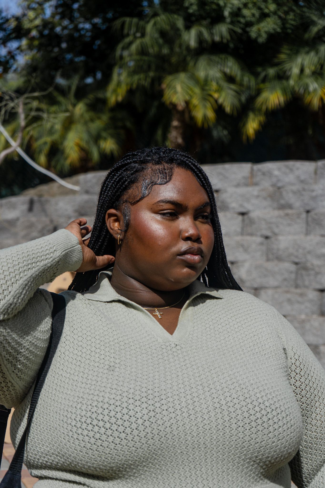 A person wearing a sage green knit sweater poses outdoors against a stone wall with palm trees in the background.