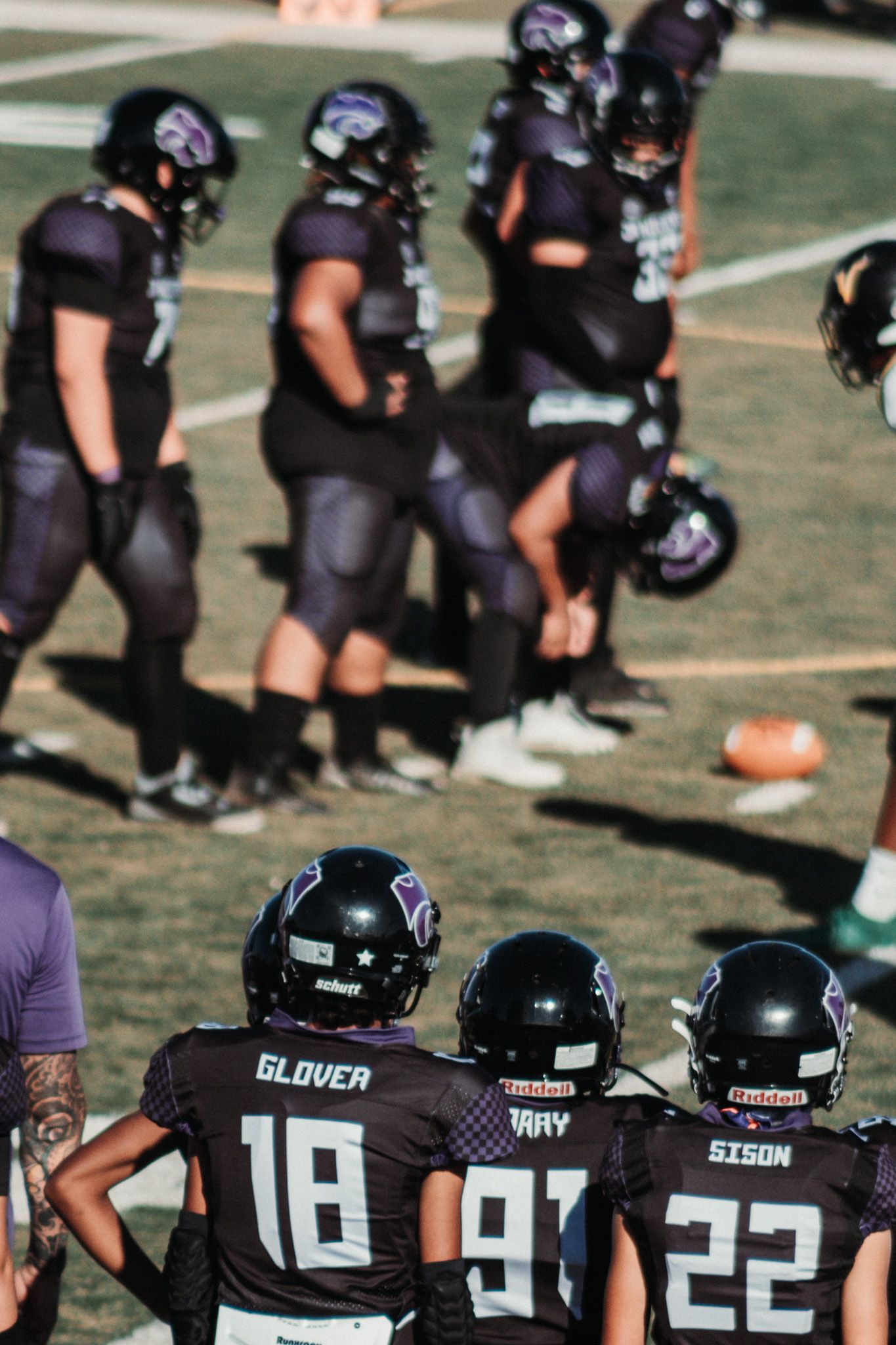 Football players in black uniforms line up on field during game while teammates watch from sideline.
