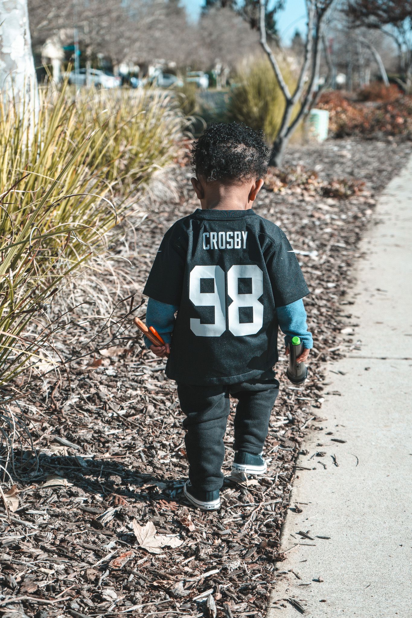 Small child wearing a black Crosby 98 sports jersey walks along a path bordered by ornamental grasses.