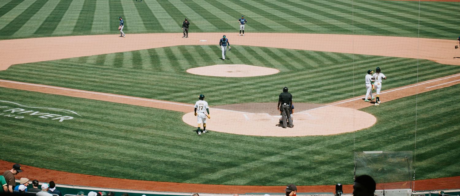 Baseball players and officials stand on a beautifully manicured diamond field with distinctive mowing patterns in the grass.