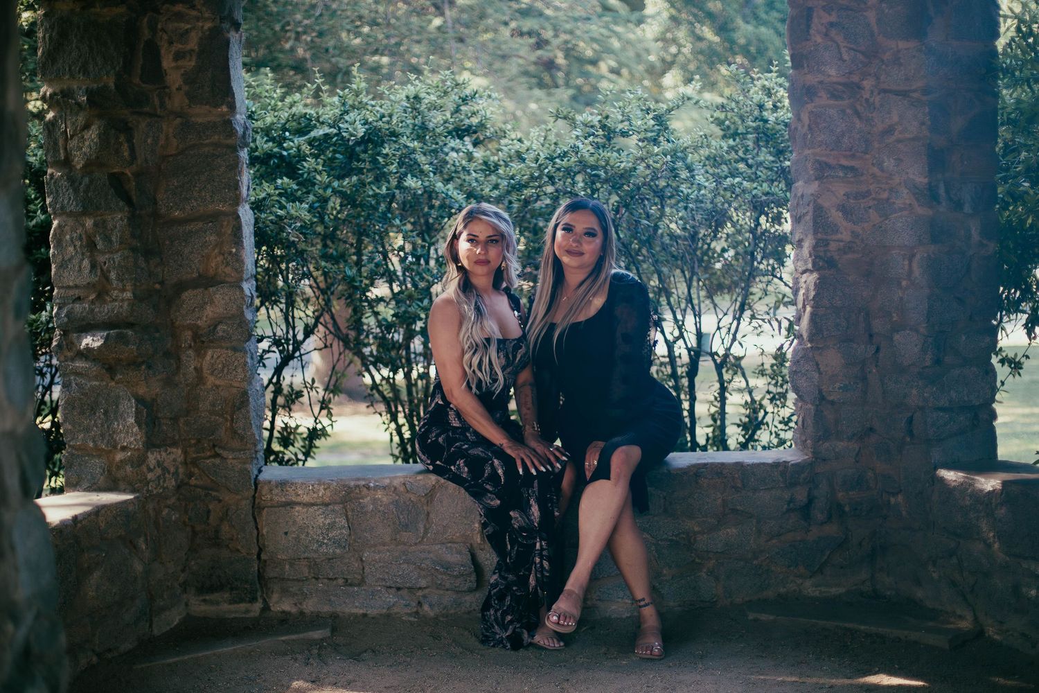 Two women pose together in a stone archway surrounded by greenery on a sunny day.