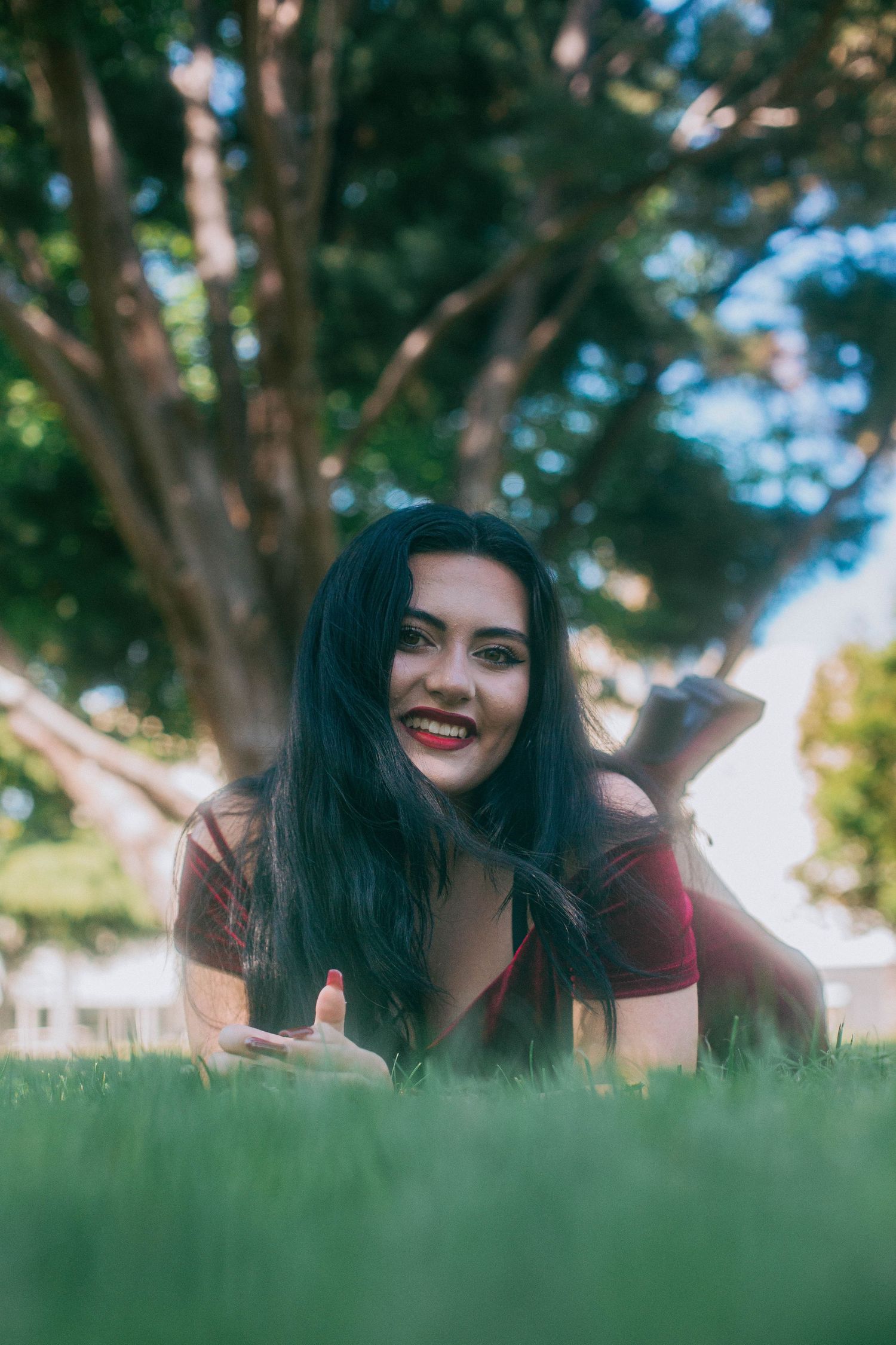 Someone lies in green grass with long dark hair wearing a burgundy top while enjoying a sunny day outdoors.