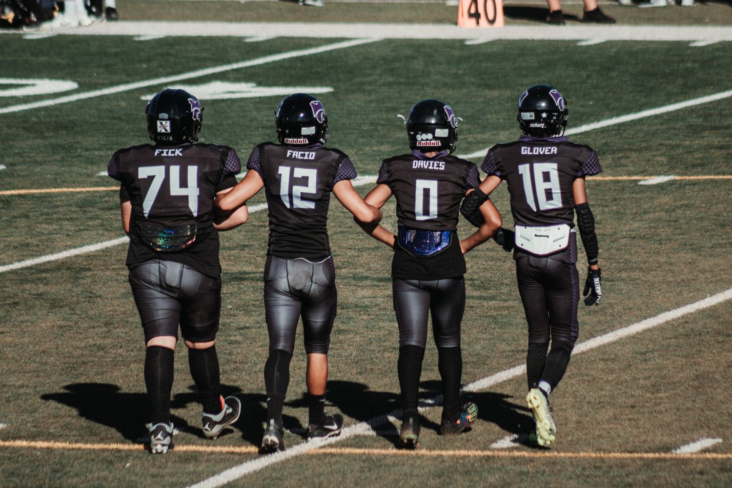 Four football players in blue and black uniforms walking together on a field with jerseys numbered 74, 12, 0, and 19.
