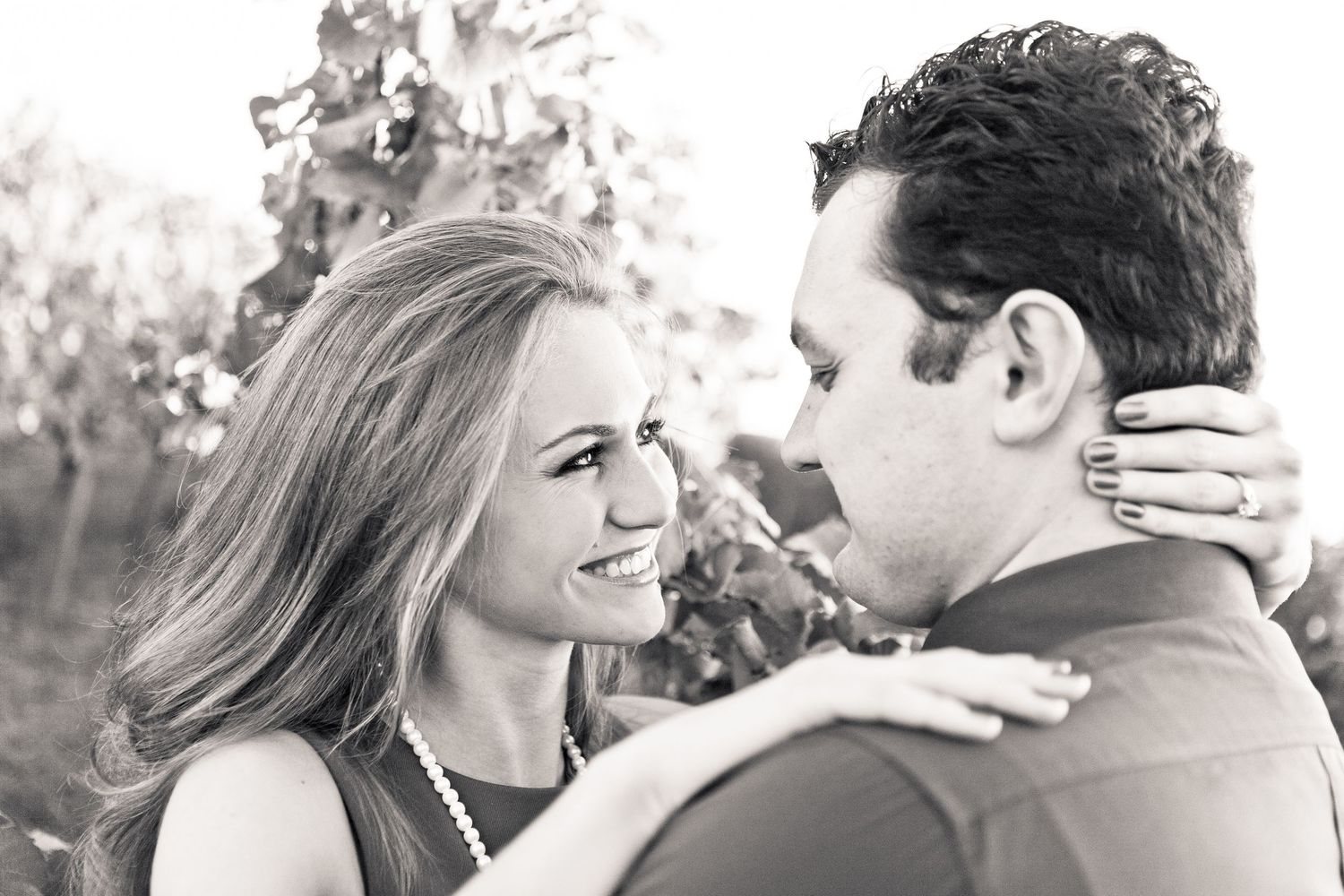 Black and white photo of a smiling couple sharing a romantic moment outdoors, woman wearing pearl necklace.