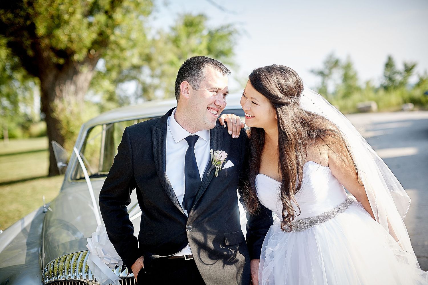 A bride and groom share a romantic moment beside a vintage white car outdoors on their wedding day.