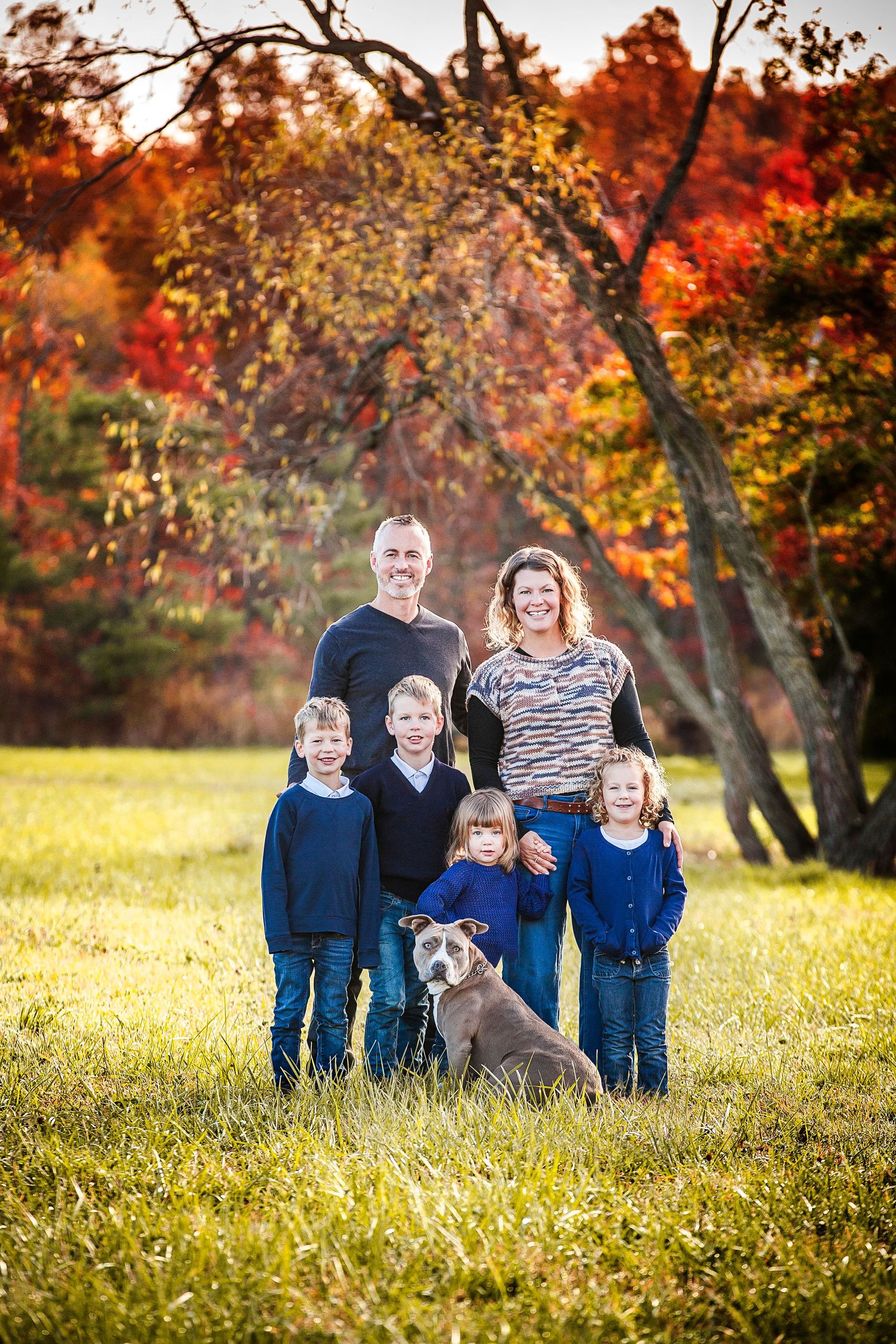 Family of six with a dog posing outdoors during fall foliage season on a sunny green lawn.