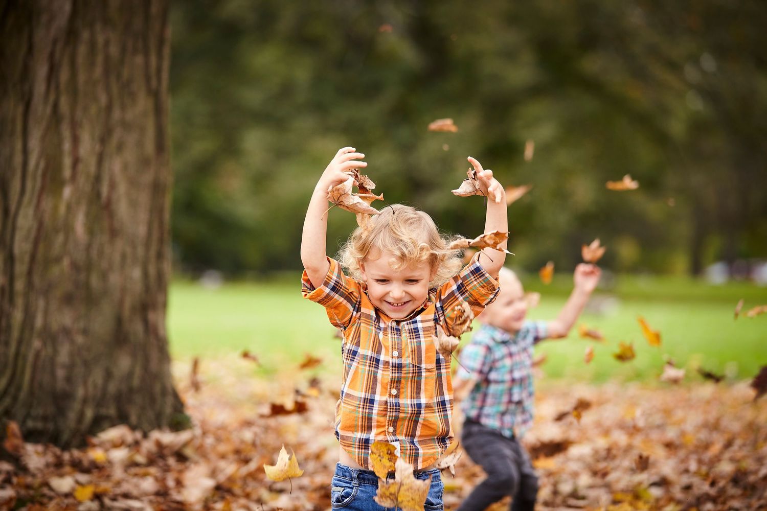 Two young children joyfully tossing autumn leaves in a park surrounded by colorful fall foliage.