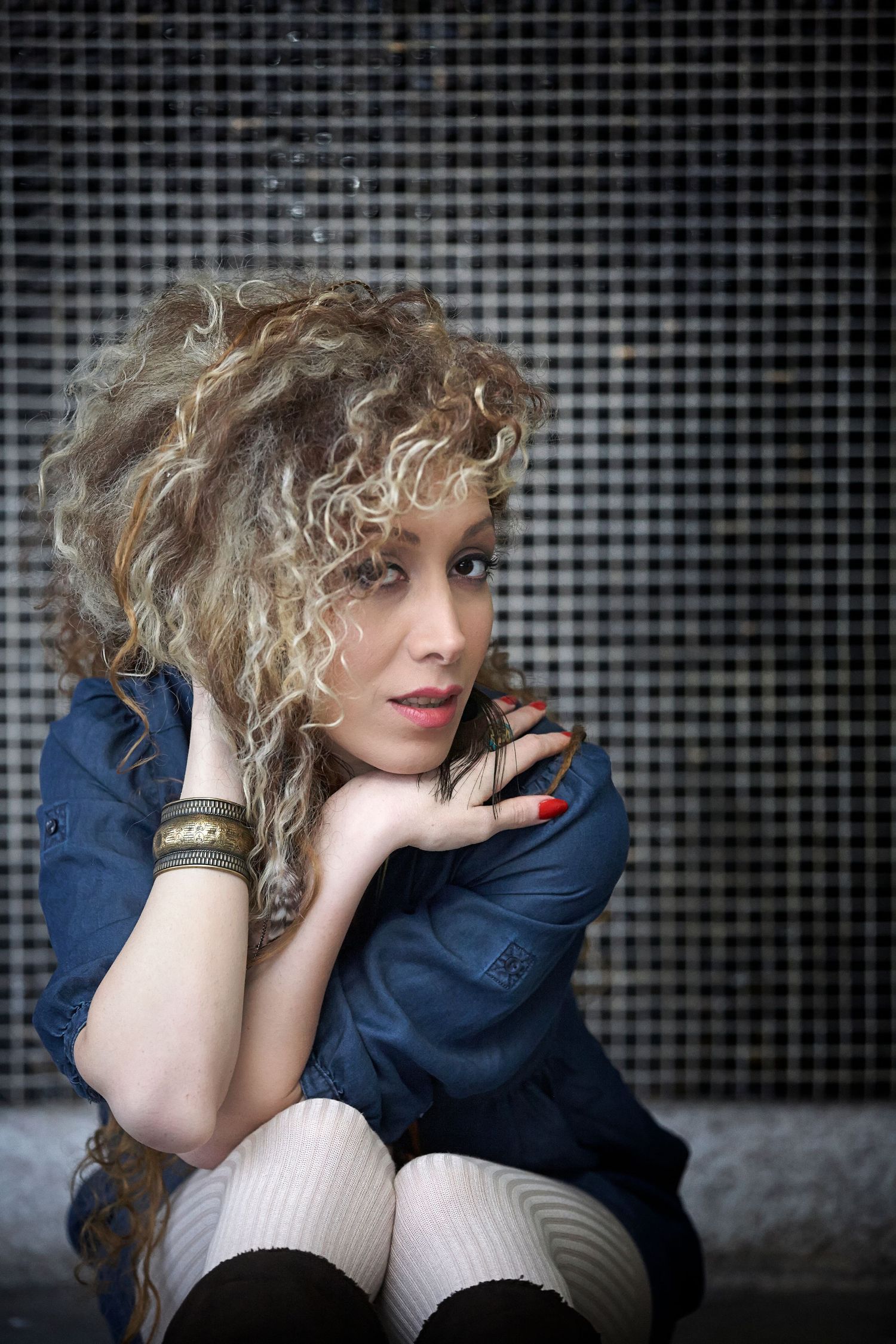 Young woman with curly blonde hair sitting against mosaic wall, wearing denim jacket and white stockings.