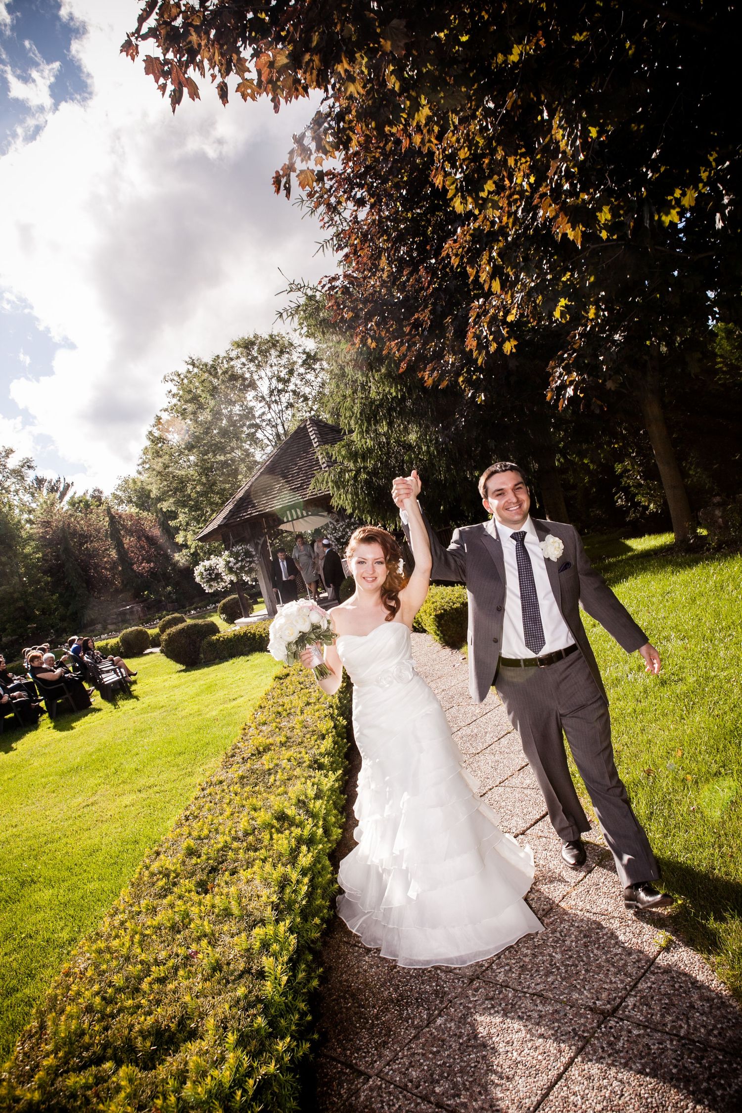 Joyful bride and groom raising hands on garden path, bride in white gown with bouquet, groom in gray suit, sunny outdoor wedding.