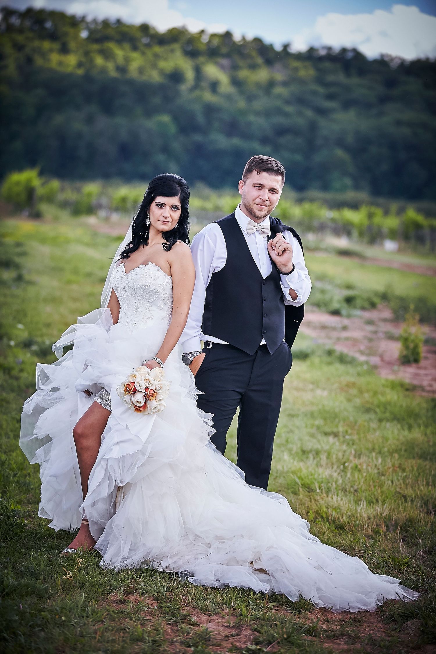 Bride in white ruffled gown holds bouquet beside groom in black suit at outdoor vineyard wedding.