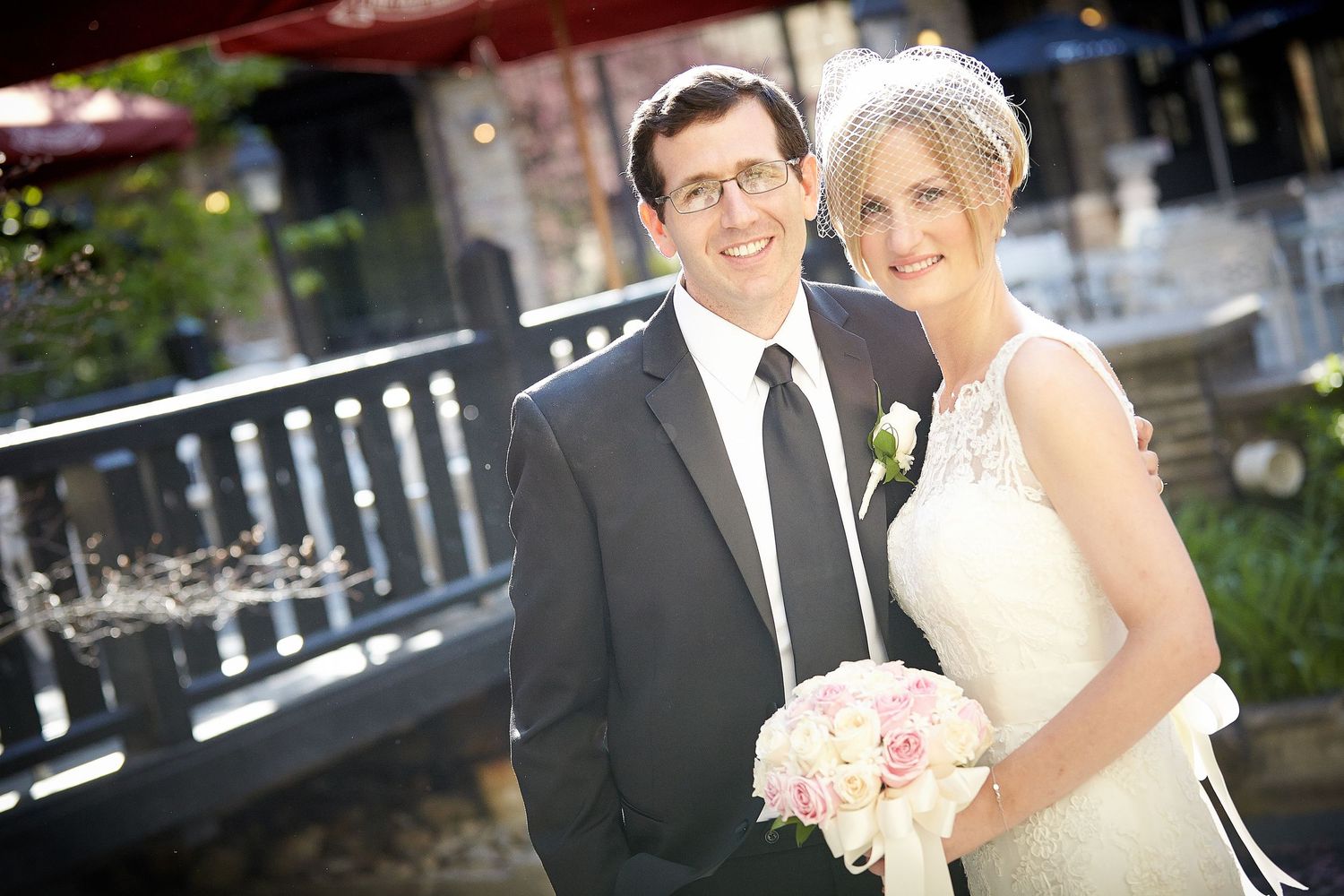 Smiling bride and groom pose together outdoors on their wedding day, bride holding pink bouquet.