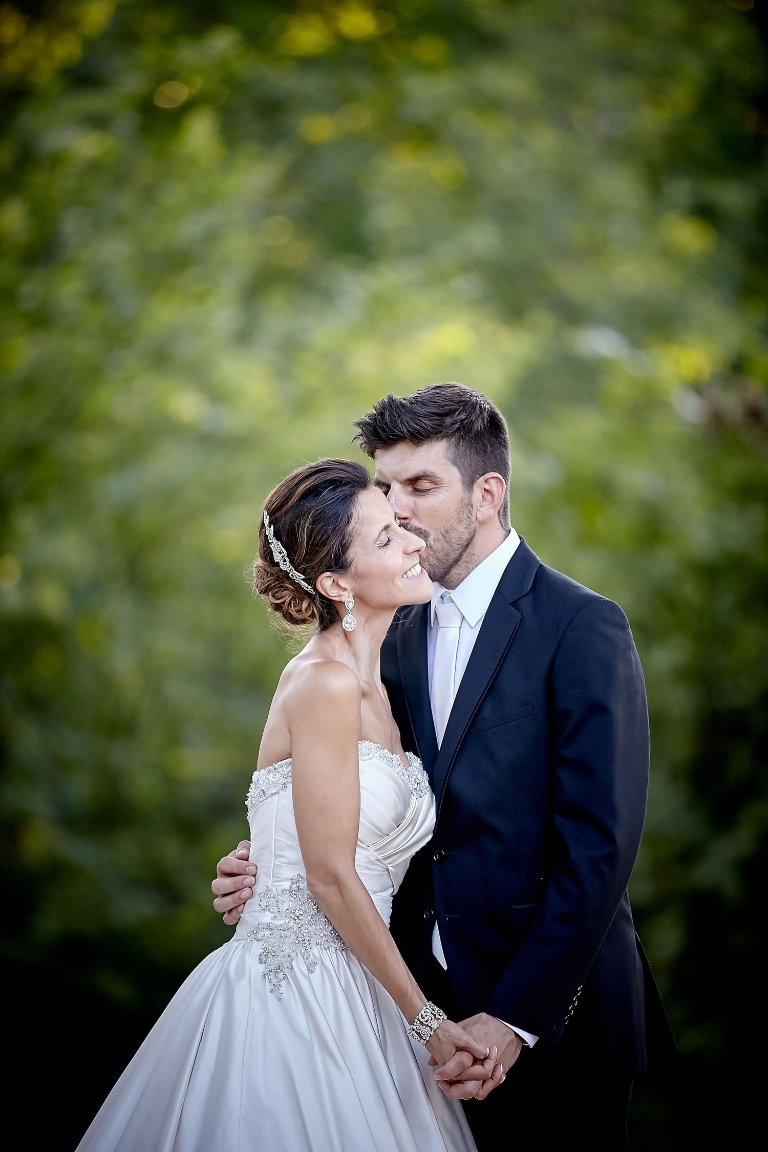 Bride and groom share a romantic kiss outdoors, surrounded by lush green trees, bride in strapless gown, groom in dark suit.