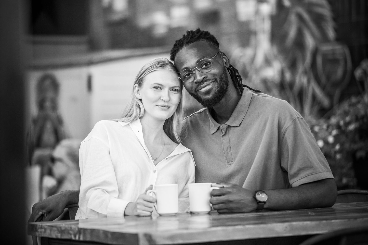 Black and white photo of a smiling interracial couple sitting together at a cafe table holding mugs.