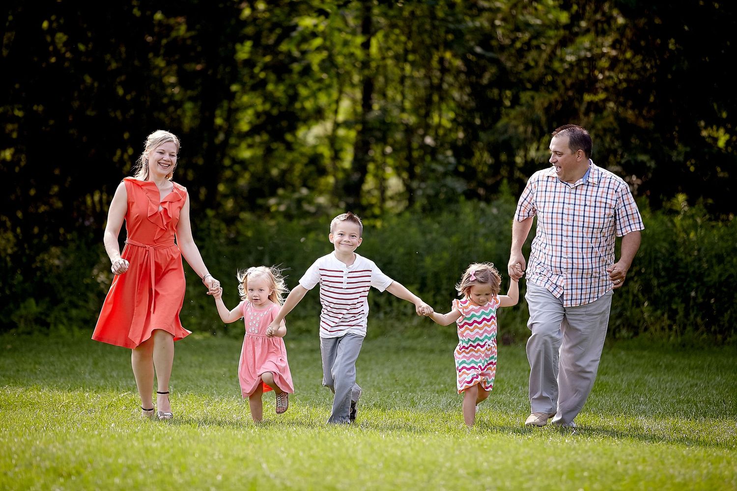 Happy family with three young children holding hands and walking together on green grass in a sunny park.
