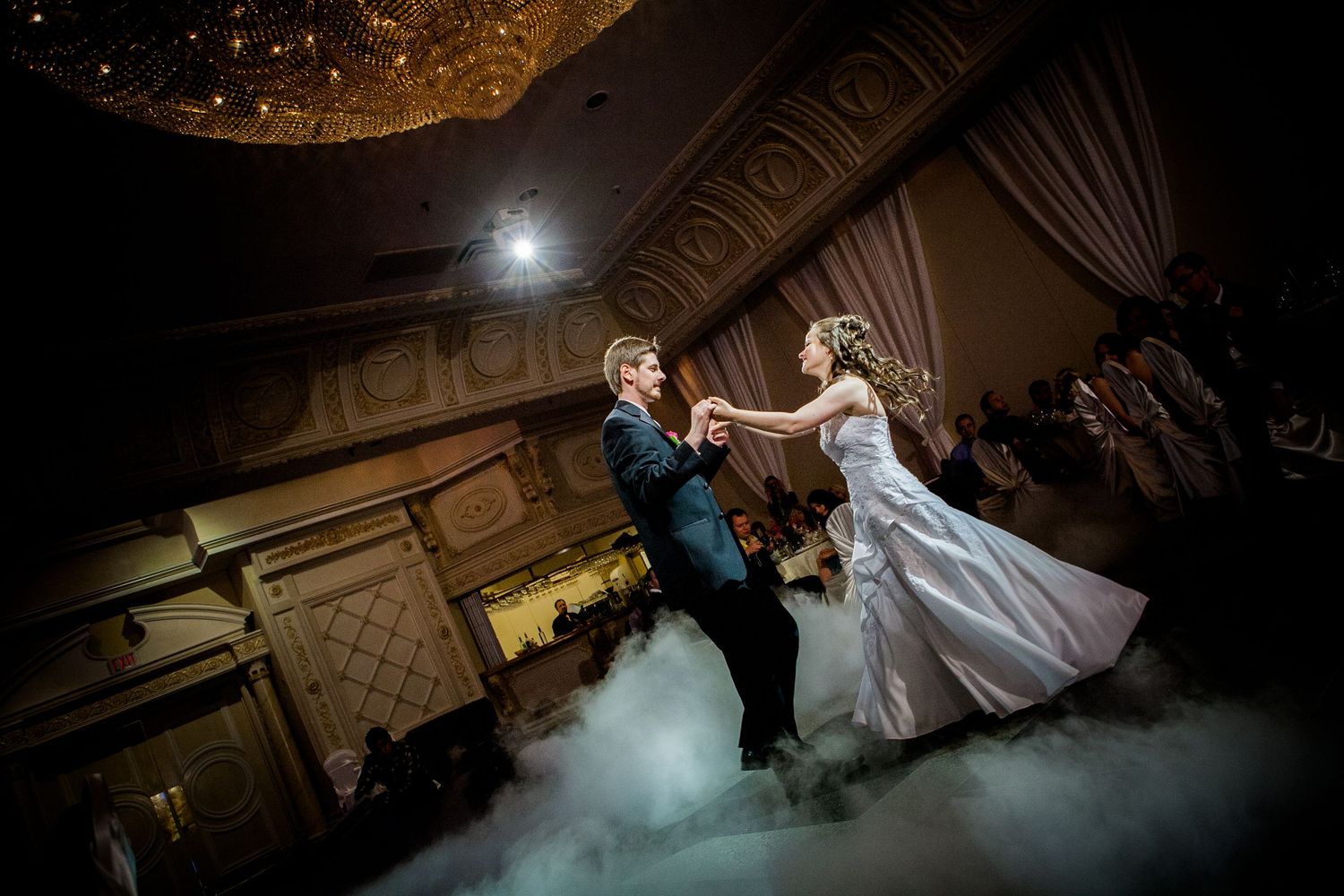 Bride and groom share first dance on fog-covered dance floor in elegant ballroom with golden chandelier.