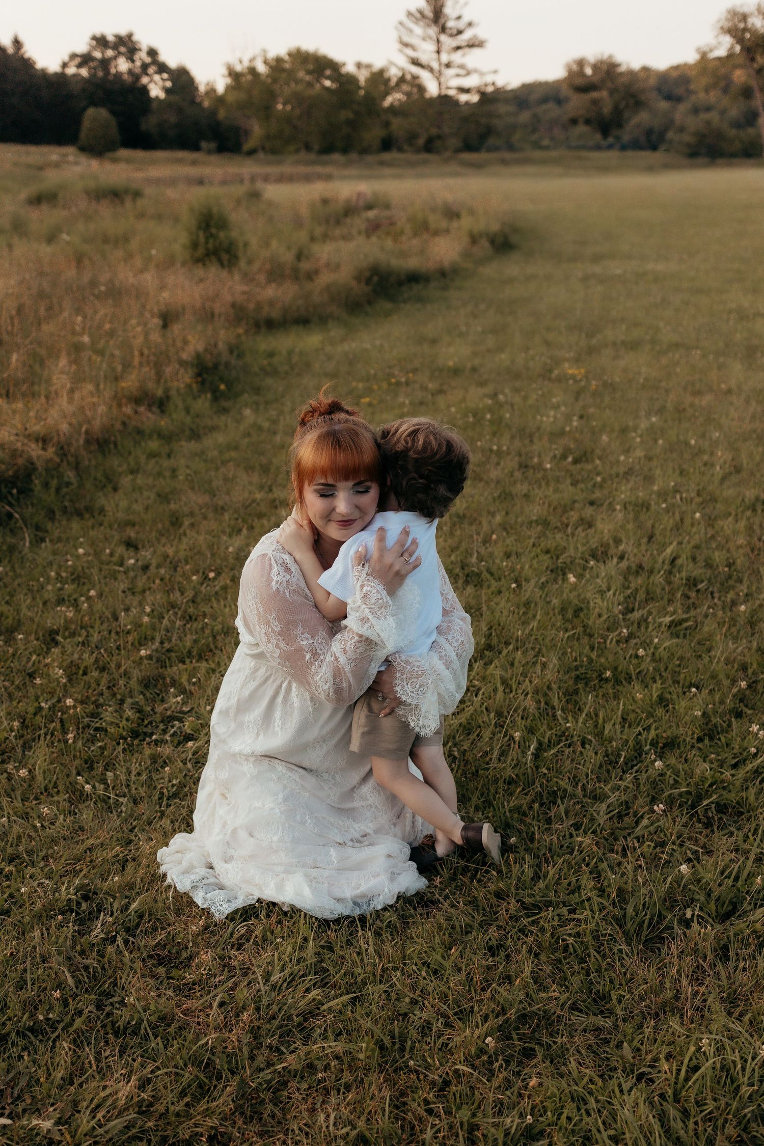 A redheaded mother in a white lace dress hugs her young son in a golden sunlit meadow.