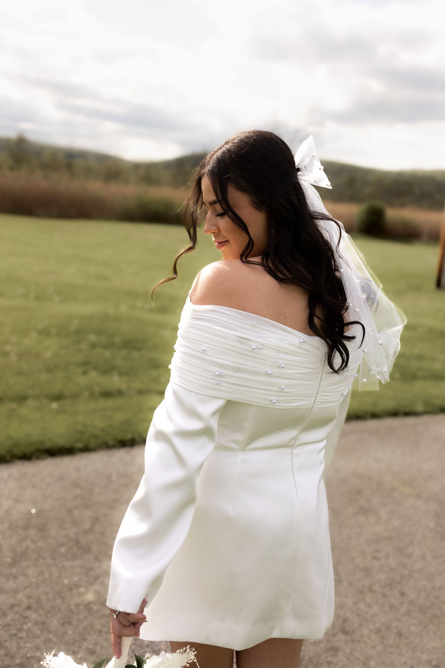 Bride in white off-shoulder mini dress with bow veil posing outdoors on her wedding day.