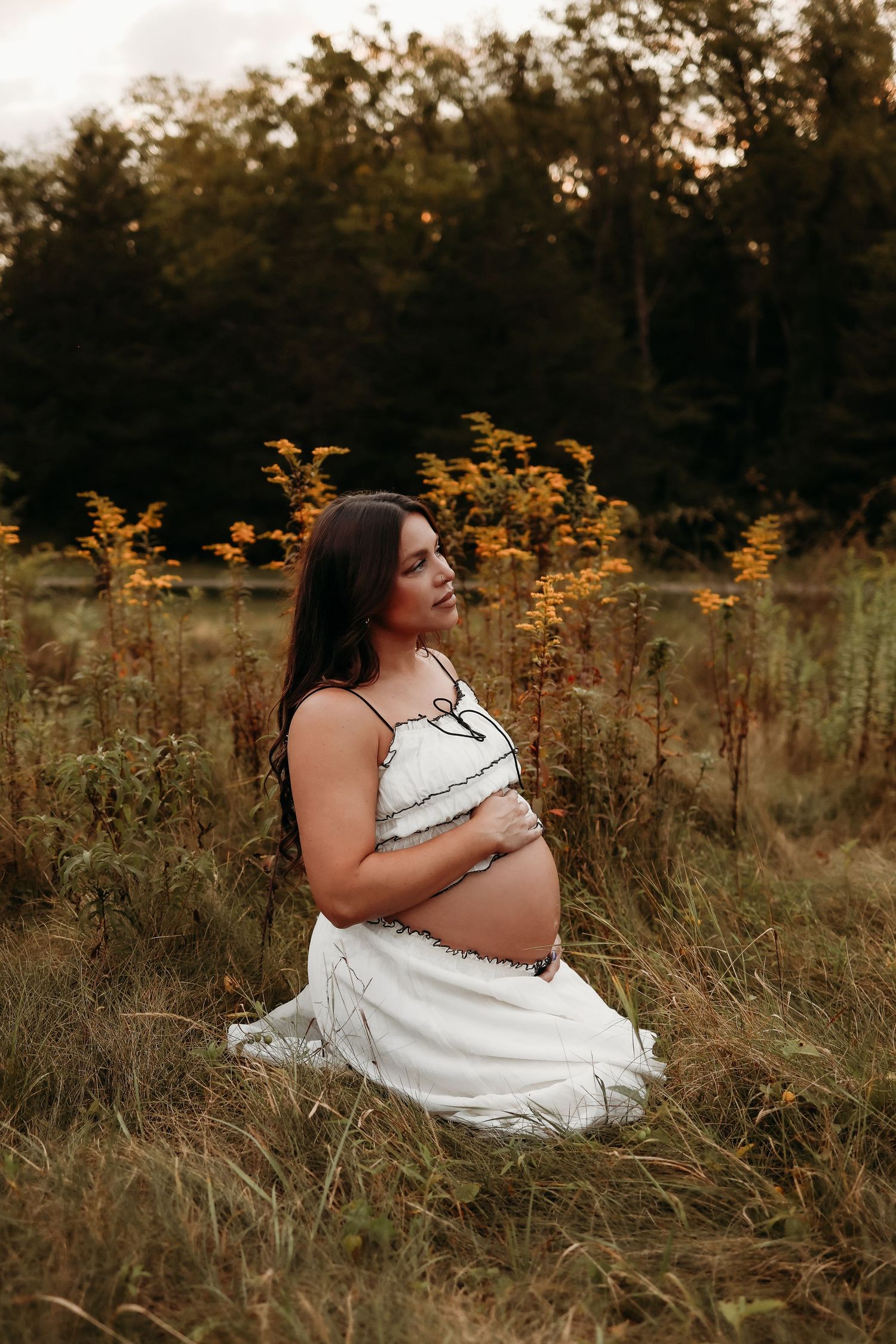 Pregnant woman in white dress kneeling in golden autumn field surrounded by wildflowers at dusk.