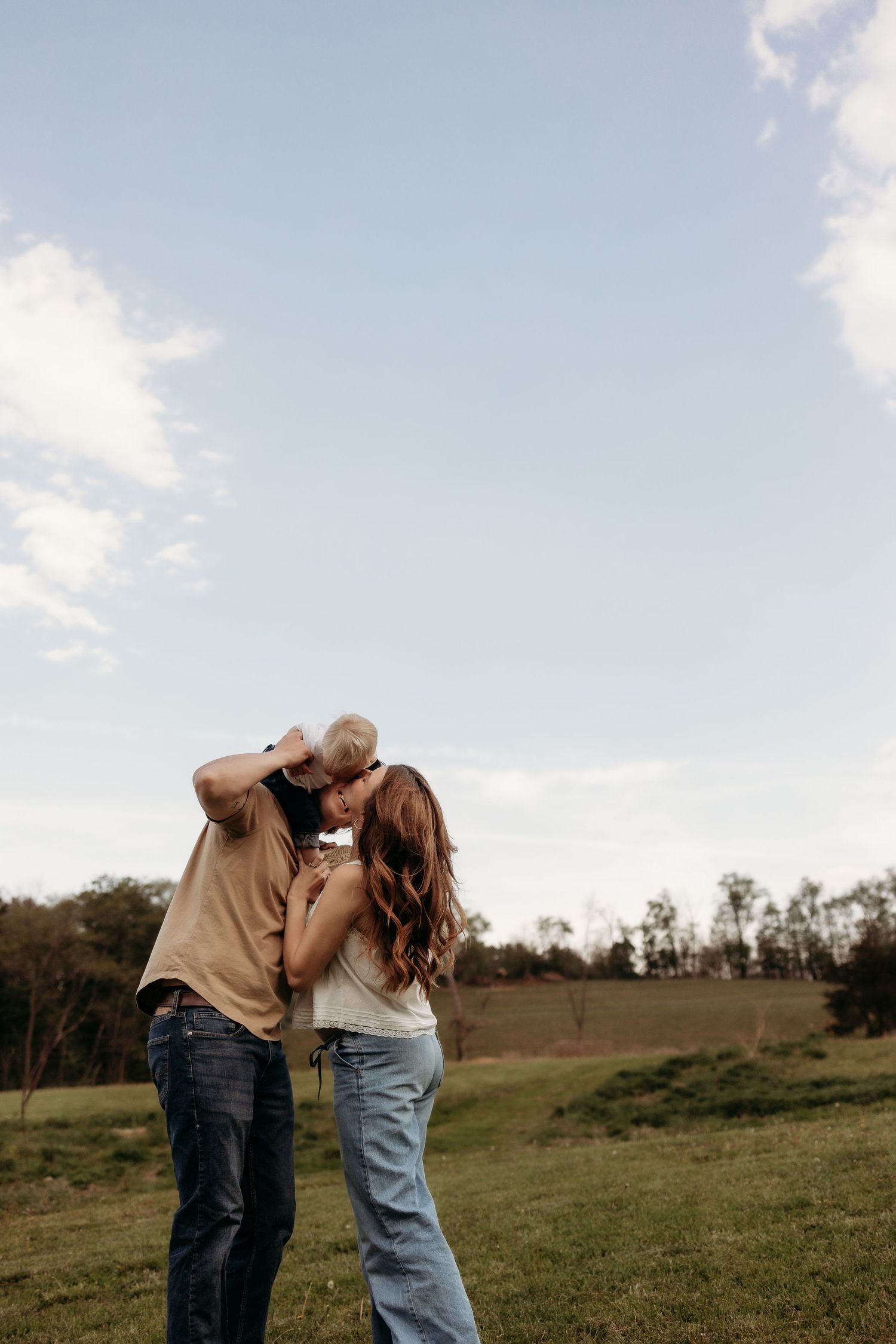A romantic couple embraces and kisses in an open green field under a partly cloudy sky.