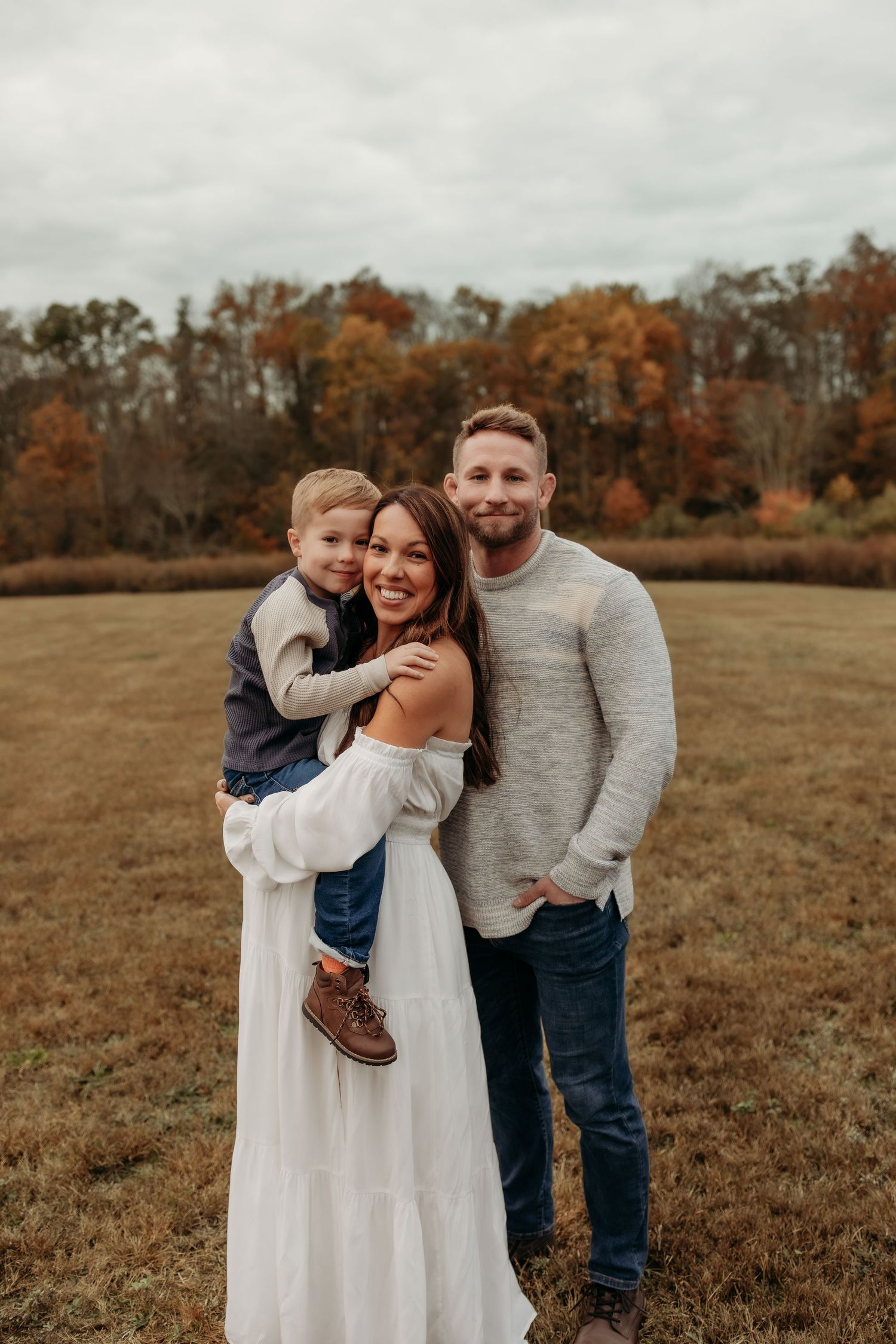 Family of three posing in fall field, mother in white dress holding son.