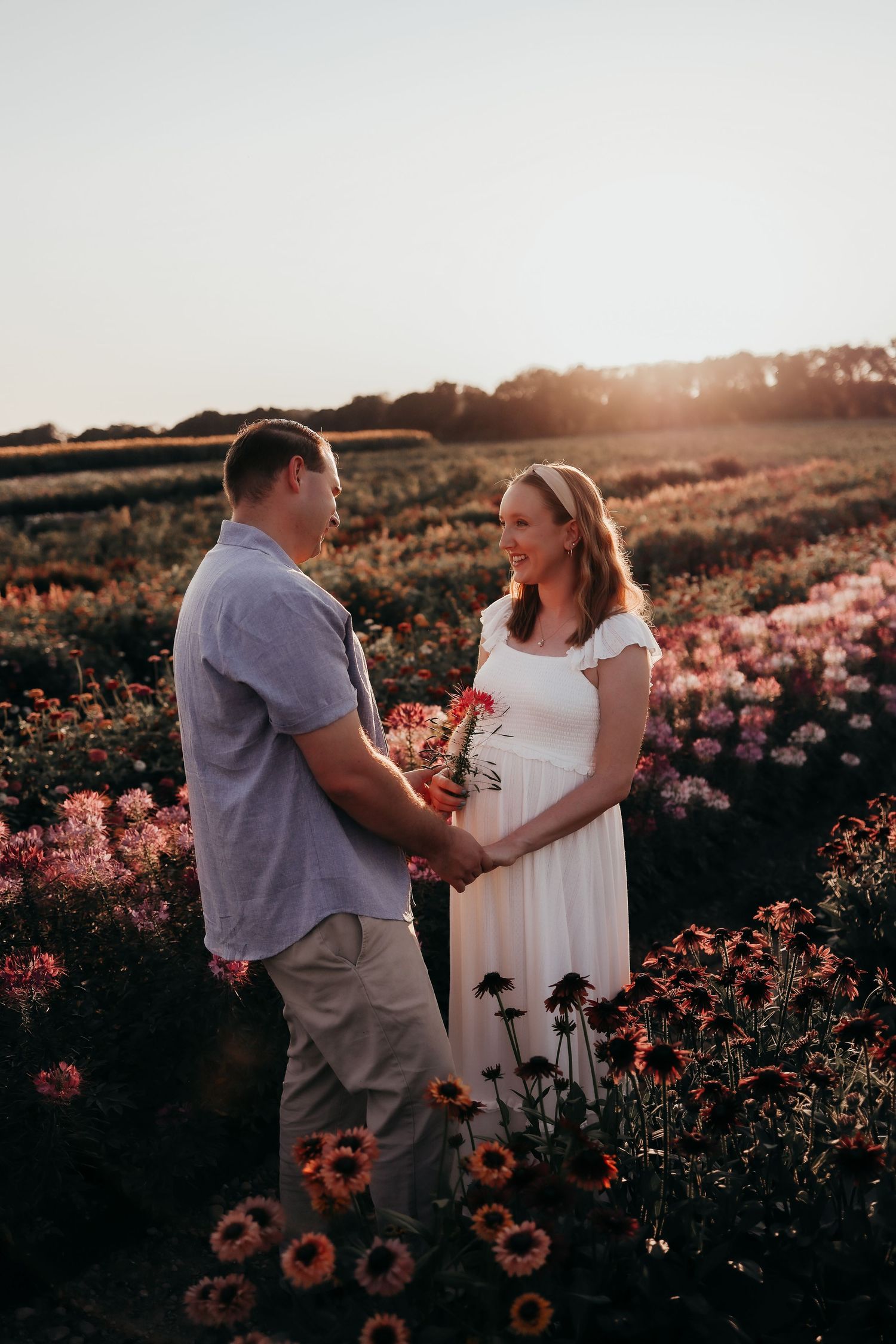 Expecting couple holding hands and laughing in a blooming flower field during a golden hour maternity shoot.