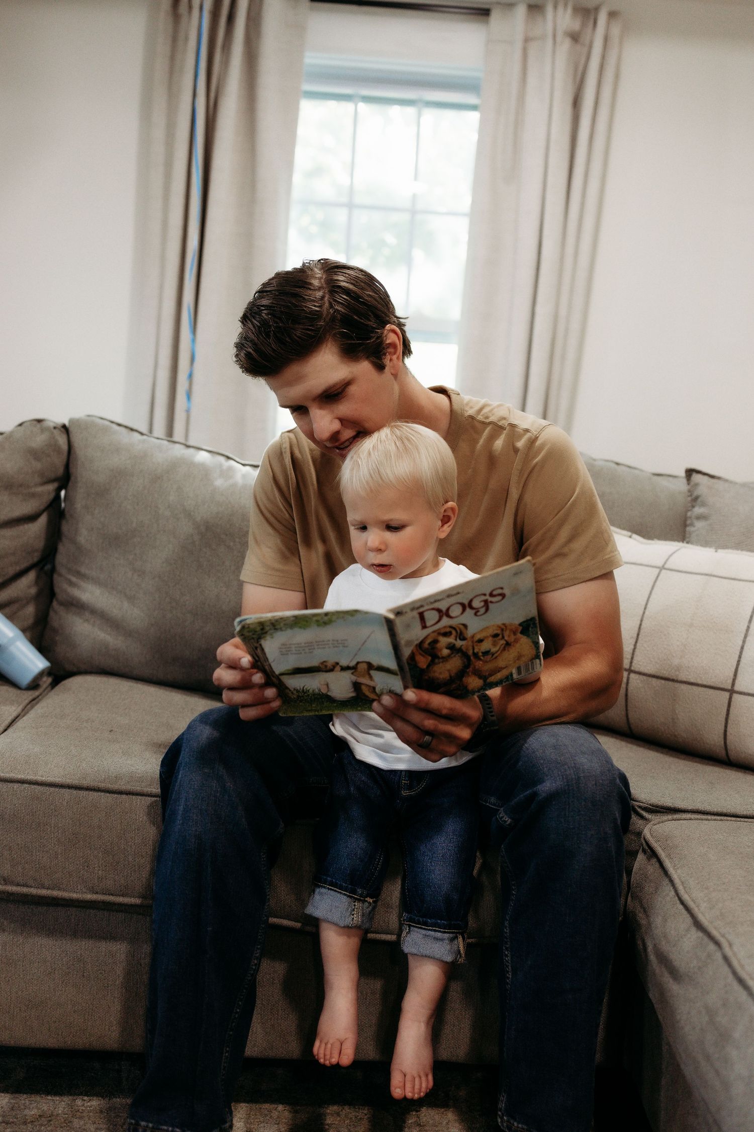 Father reading a Dogs book with his toddler son on a cozy gray sectional sofa.