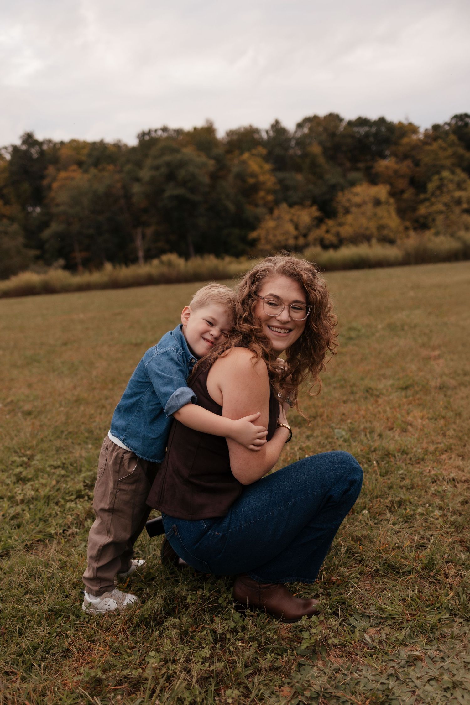 A smiling woman with curly hair and glasses crouches as a young boy hugs her from behind in an autumn field.