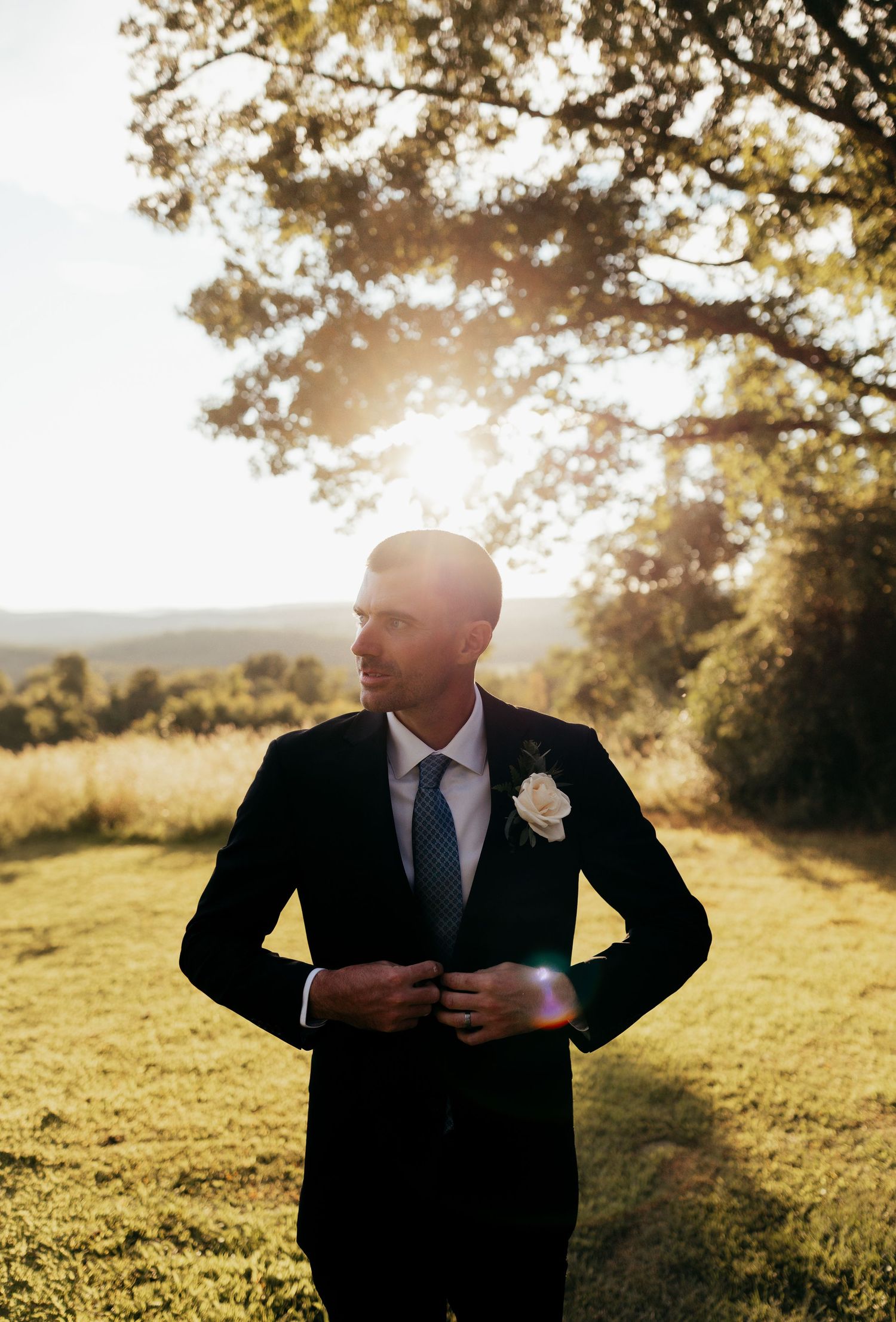 Groom in black suit with white boutonniere adjusting jacket outdoors at golden hour with sunlight through trees.