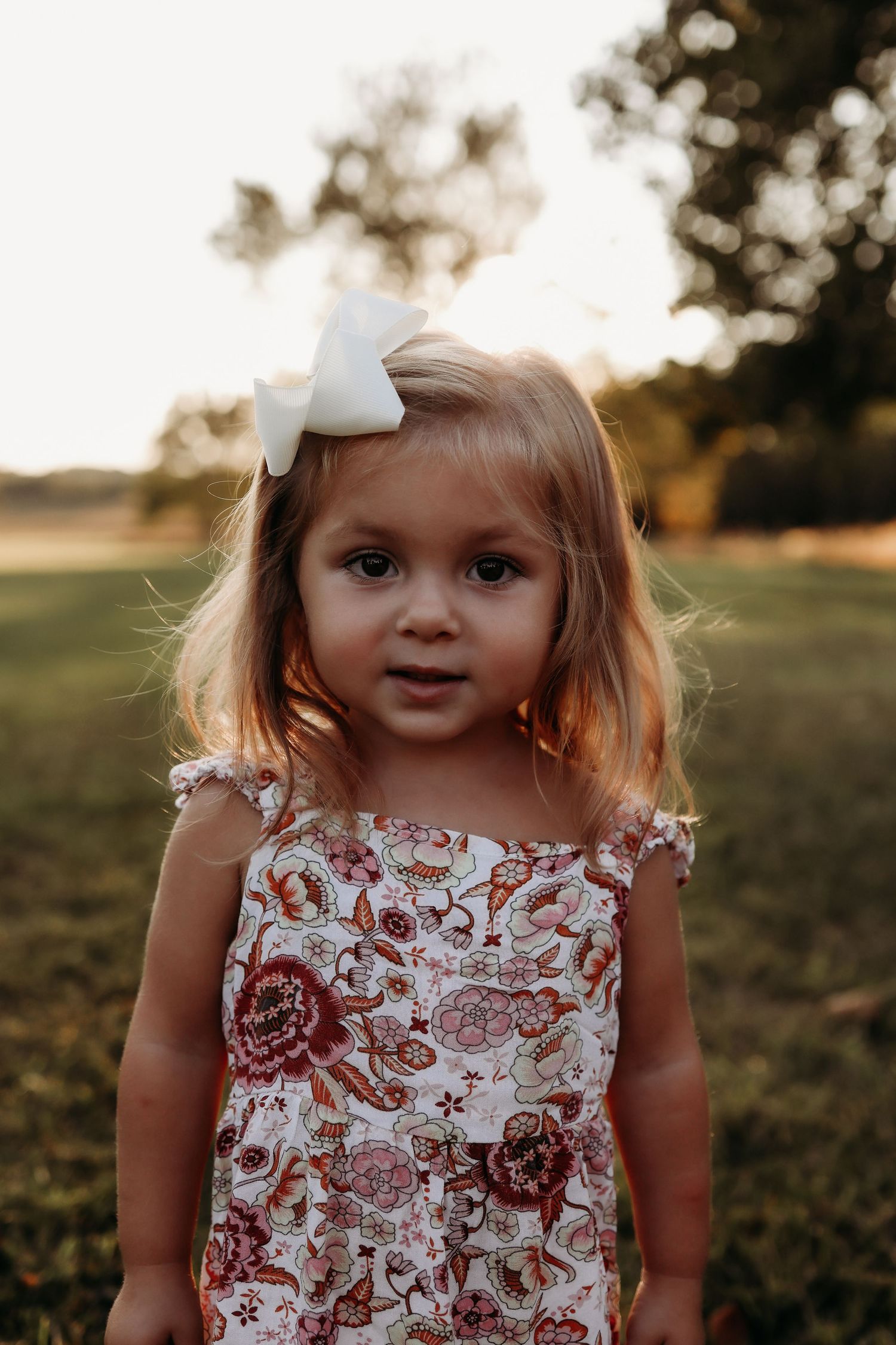 Adorable toddler girl in floral dress with white bow hair clip poses outdoors during golden hour sunset.