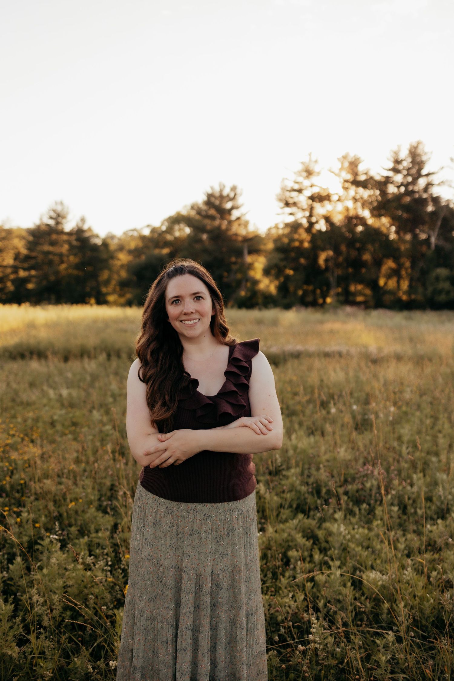 Smiling woman with arms crossed stands in golden meadow with tall grass and sunlit trees in the background.