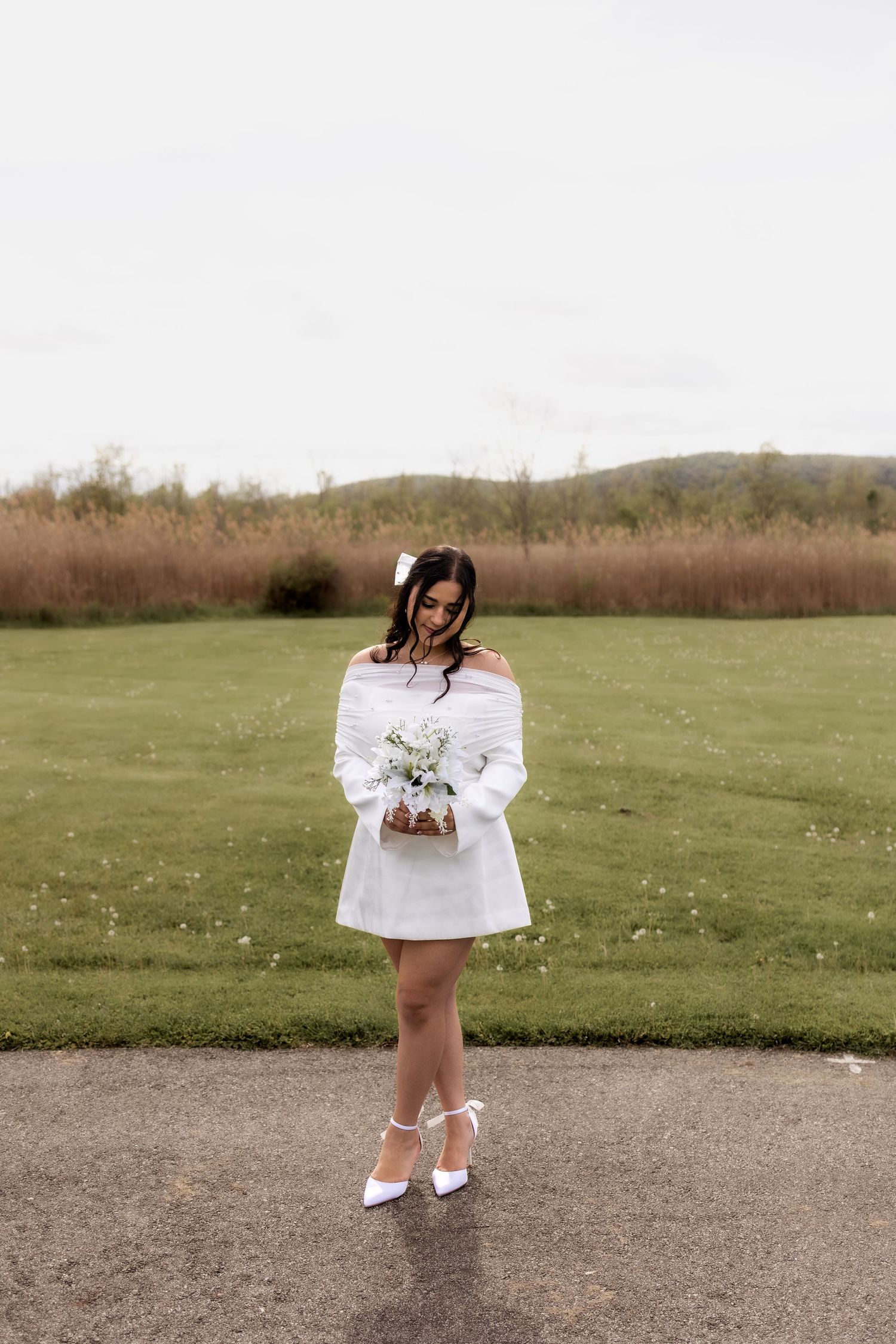 Bride in white off-shoulder mini dress holding a small floral bouquet standing on a path in a green field.