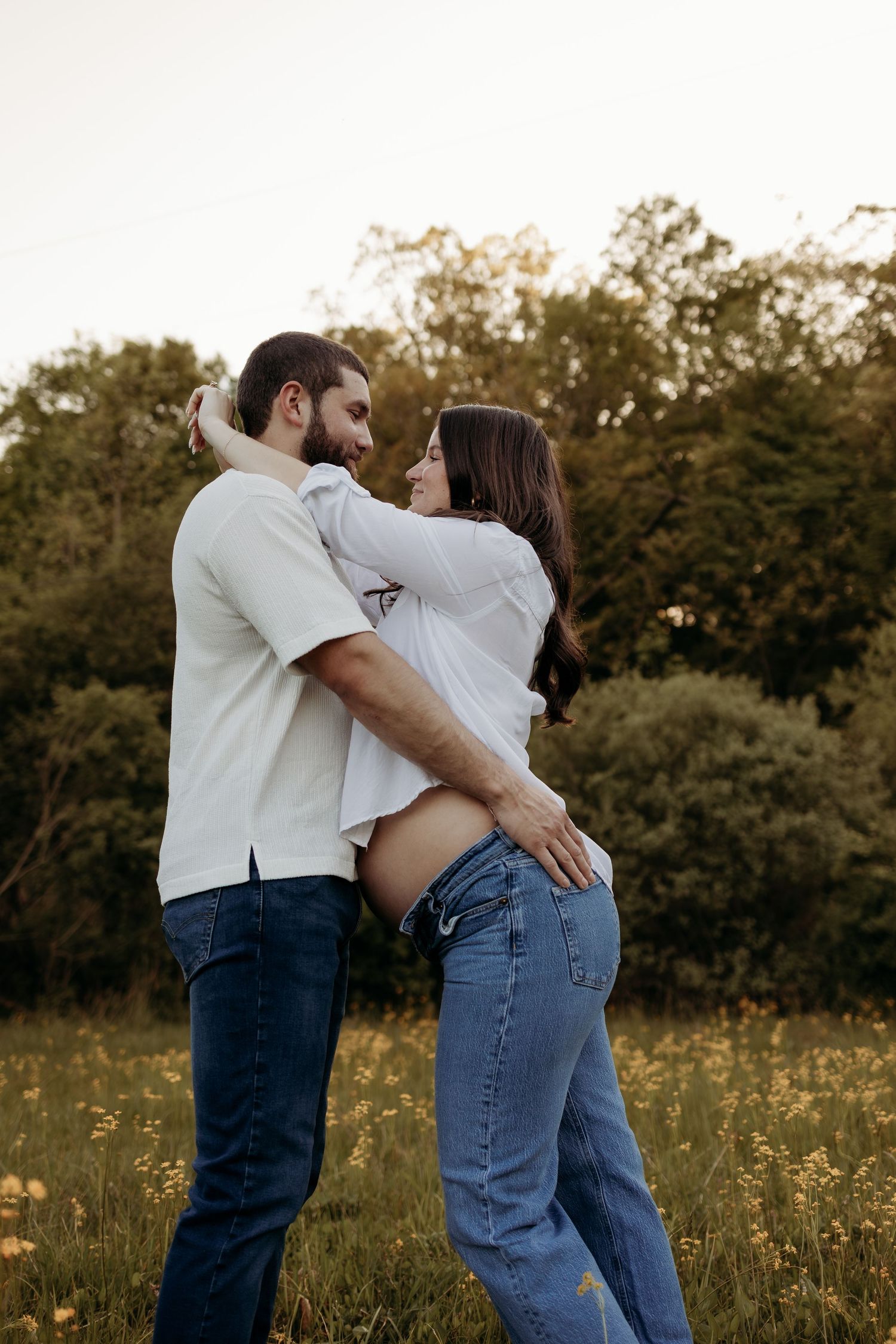 Expecting couple embraces face to face in sunlit field surrounded by wildflowers and green trees.