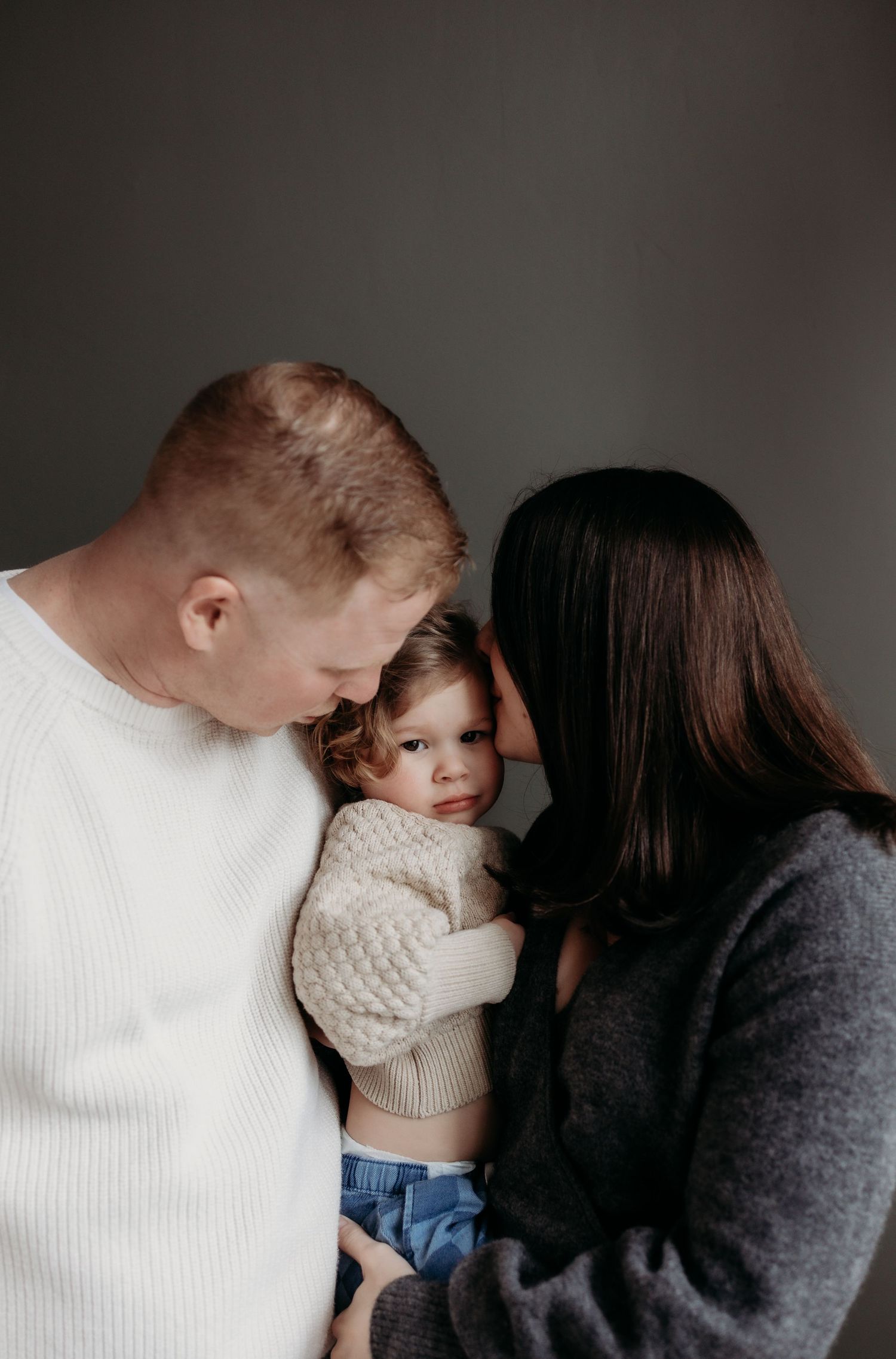 Parents tenderly holding their toddler child in a warm indoor family portrait.