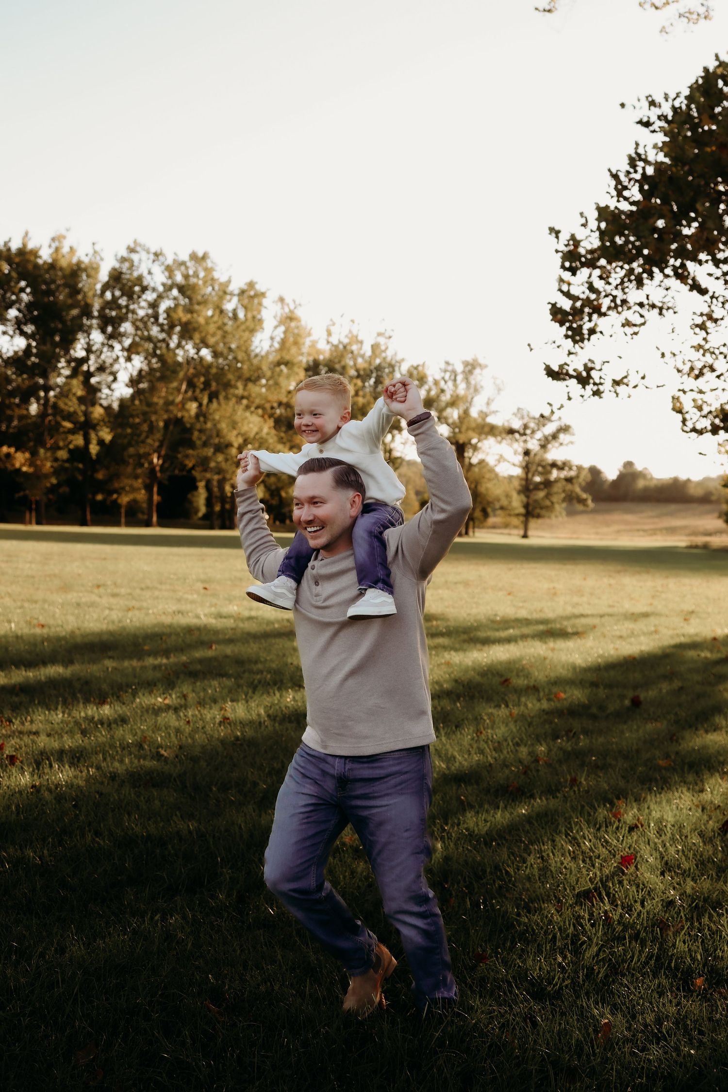 Father laughing while carrying young child on shoulders in sunny park during golden hour.