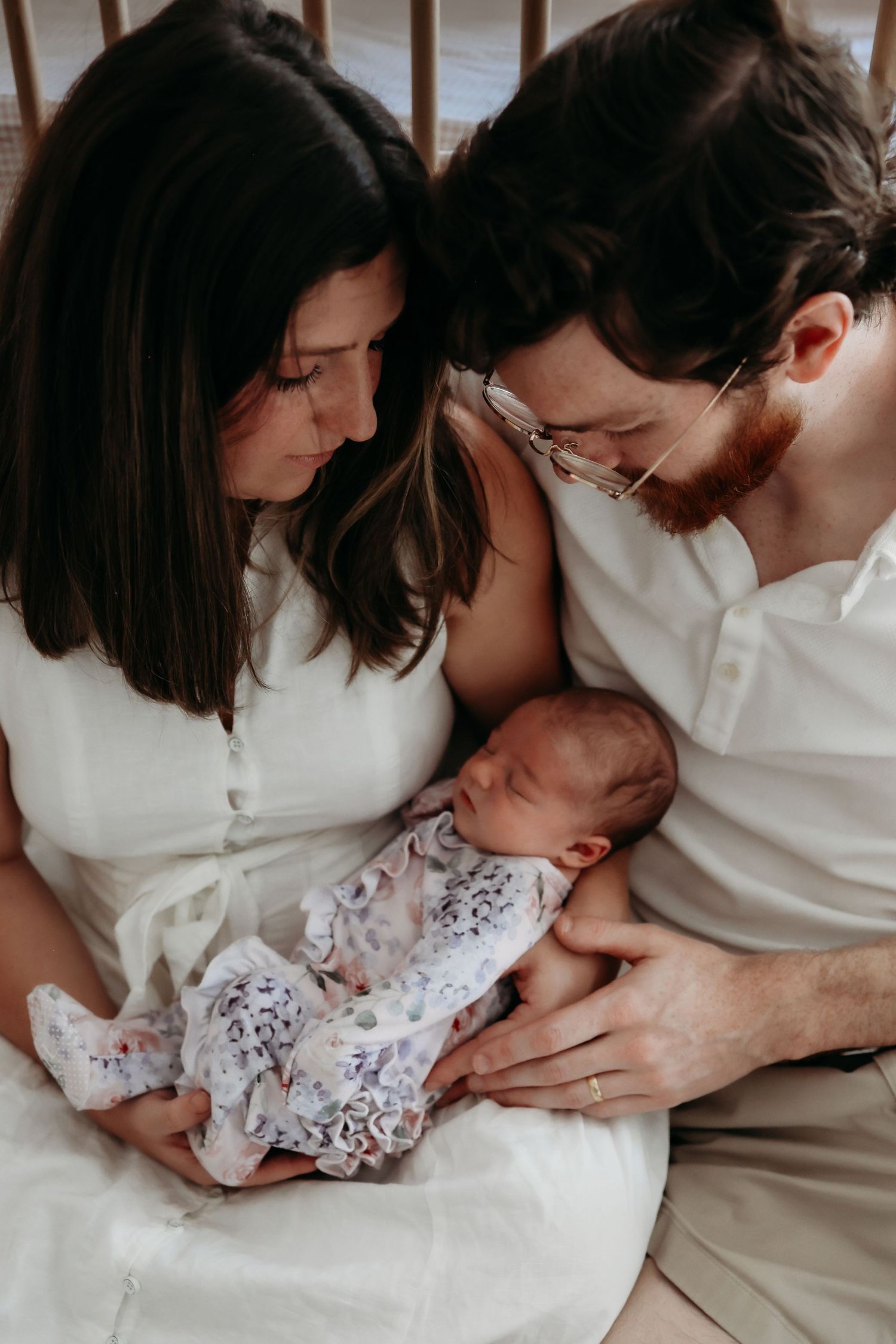 New parents in white outfits lovingly gaze down at sleeping newborn baby in their arms.