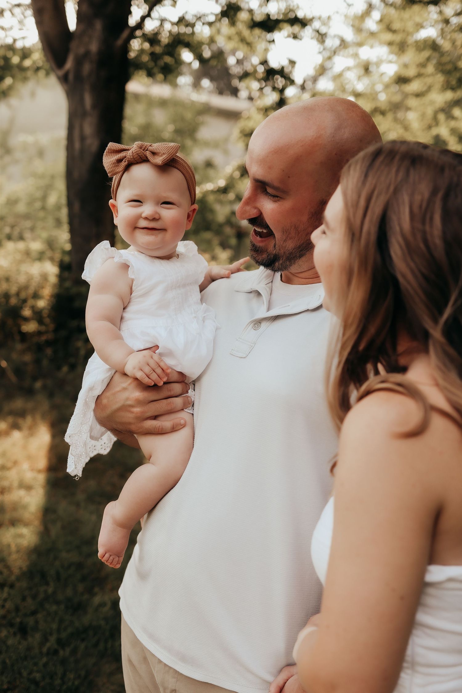 Happy bald father holding smiling baby girl in white dress while mother looks on outdoors.