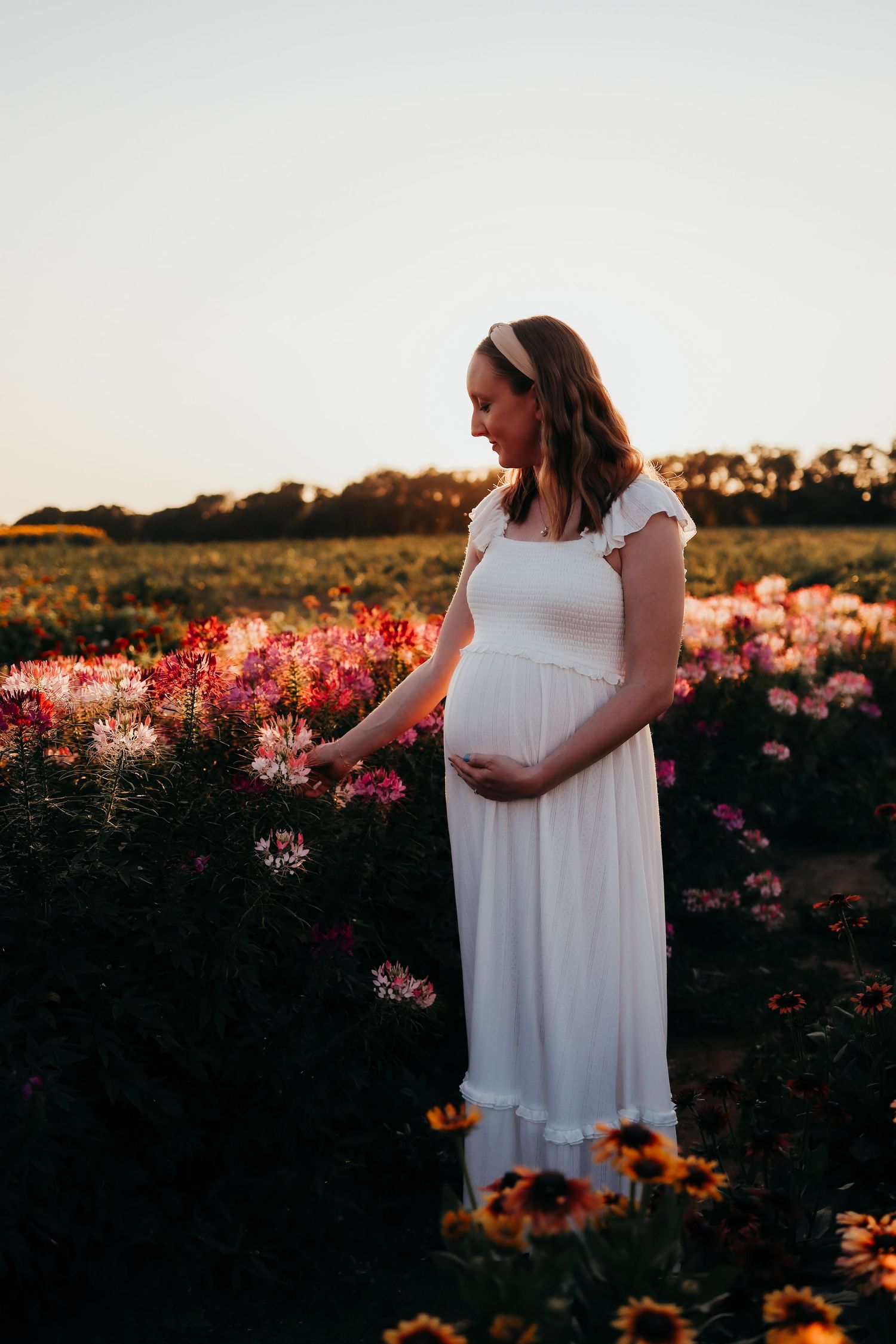 A woman in a flowing white dress stands among vibrant pink and orange flowers at sunset for a maternity shoot in Newton, New Jersey
