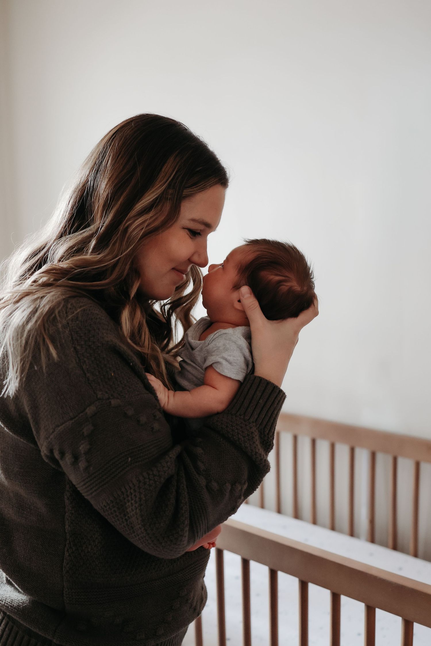 A mother tenderly holds her newborn baby close to her face near a wooden crib.