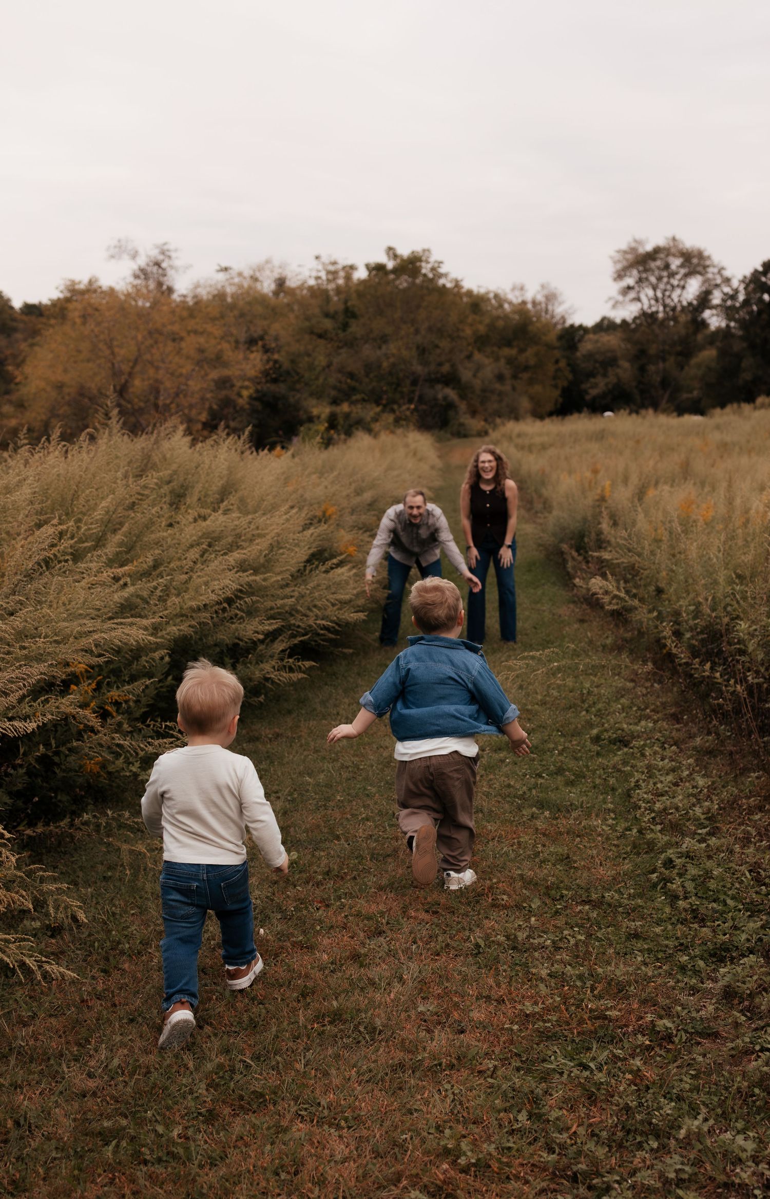 Two young children run toward their parents along a grassy path surrounded by tall golden meadow grass in an autumn outdoor setting.