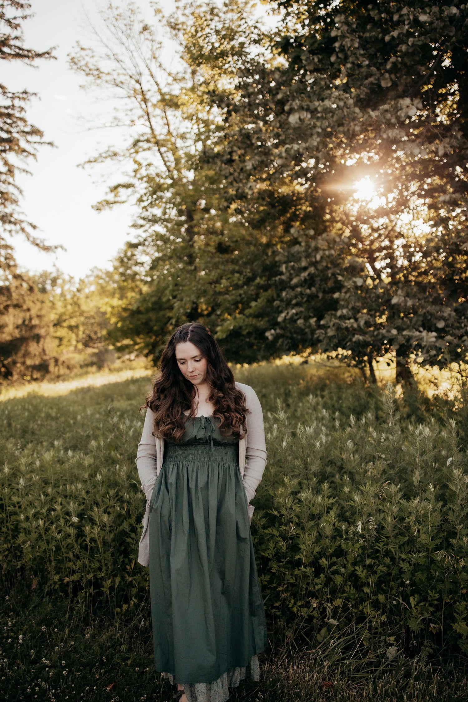 Young woman in green dress and cardigan stands in sunlit meadow surrounded by blooming trees at golden hour.
