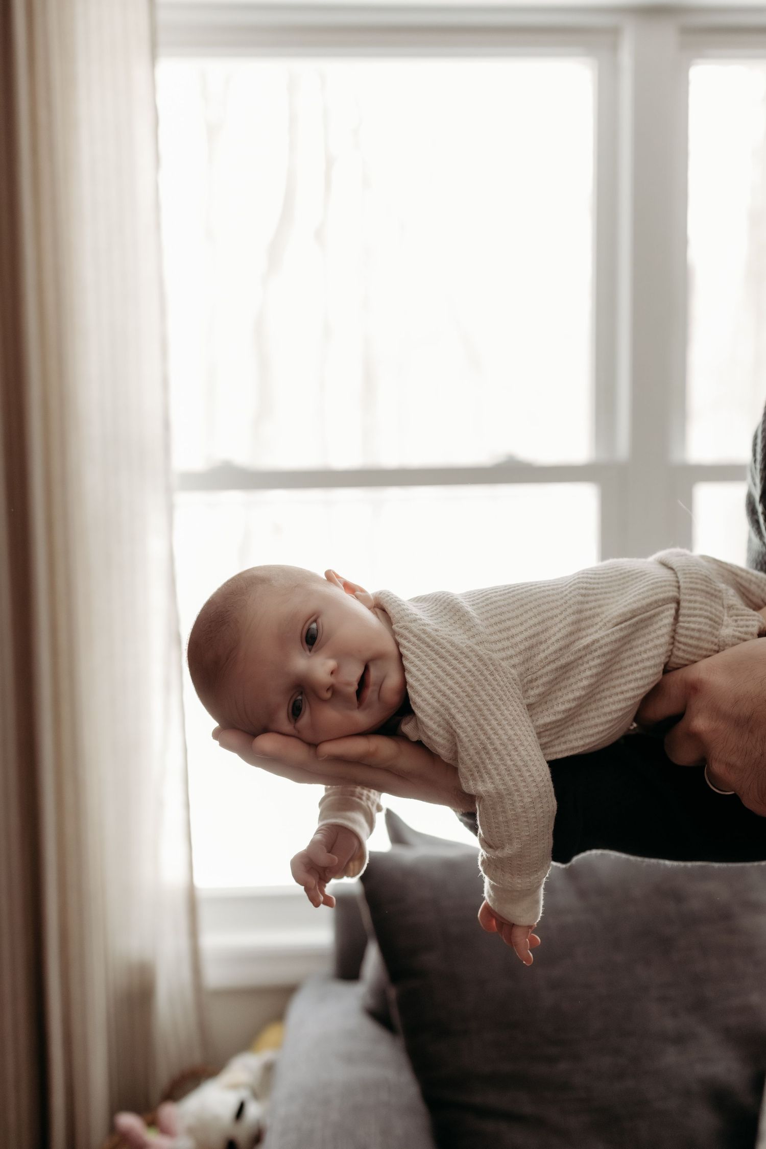 Alert newborn baby in striped onesie being held horizontally near bright window.