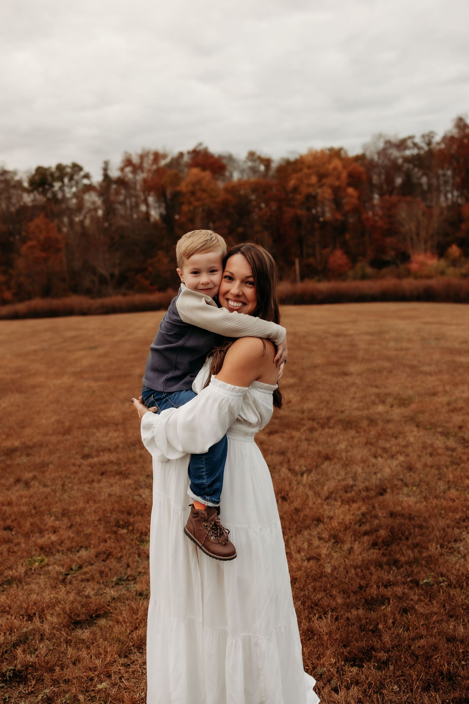 A mother in a flowing white dress holds a small child in an autumn field with orange-tinted trees in the background.