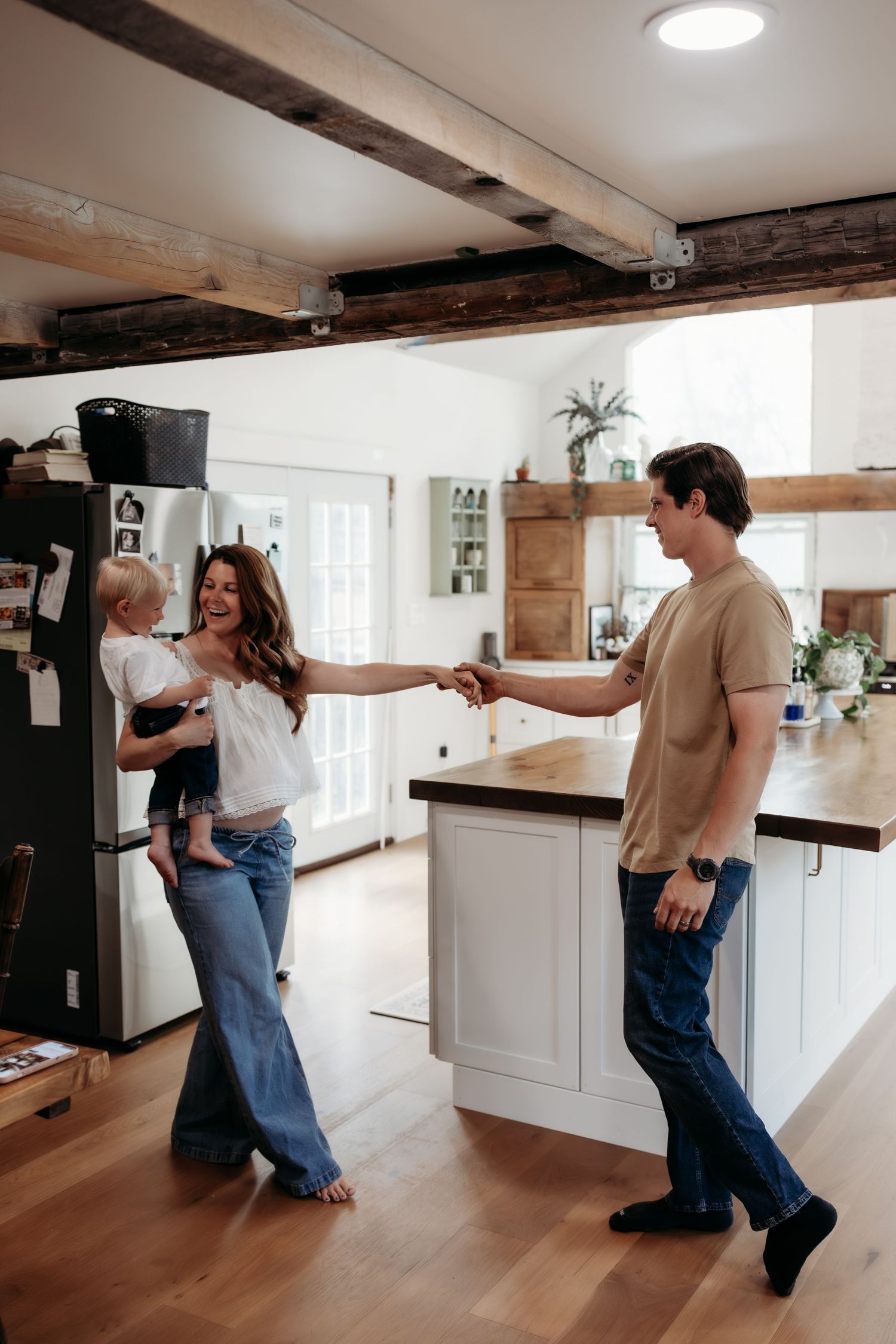 A couple dances in a rustic kitchen while the woman holds a toddler, all wearing casual jeans in a bright home.